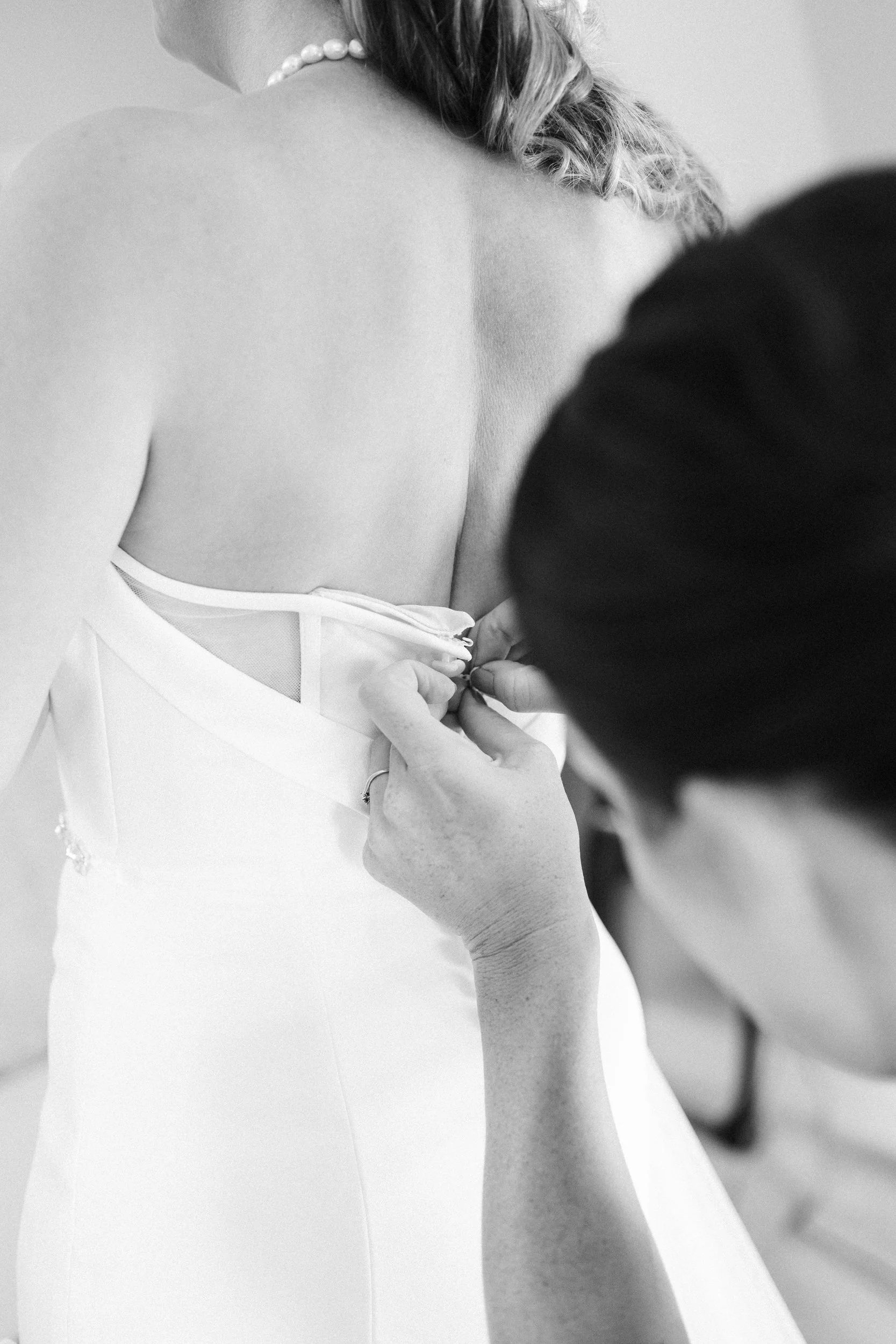 A woman is being helped into her wedding dress by another person, who is adjusting the back of the gown.