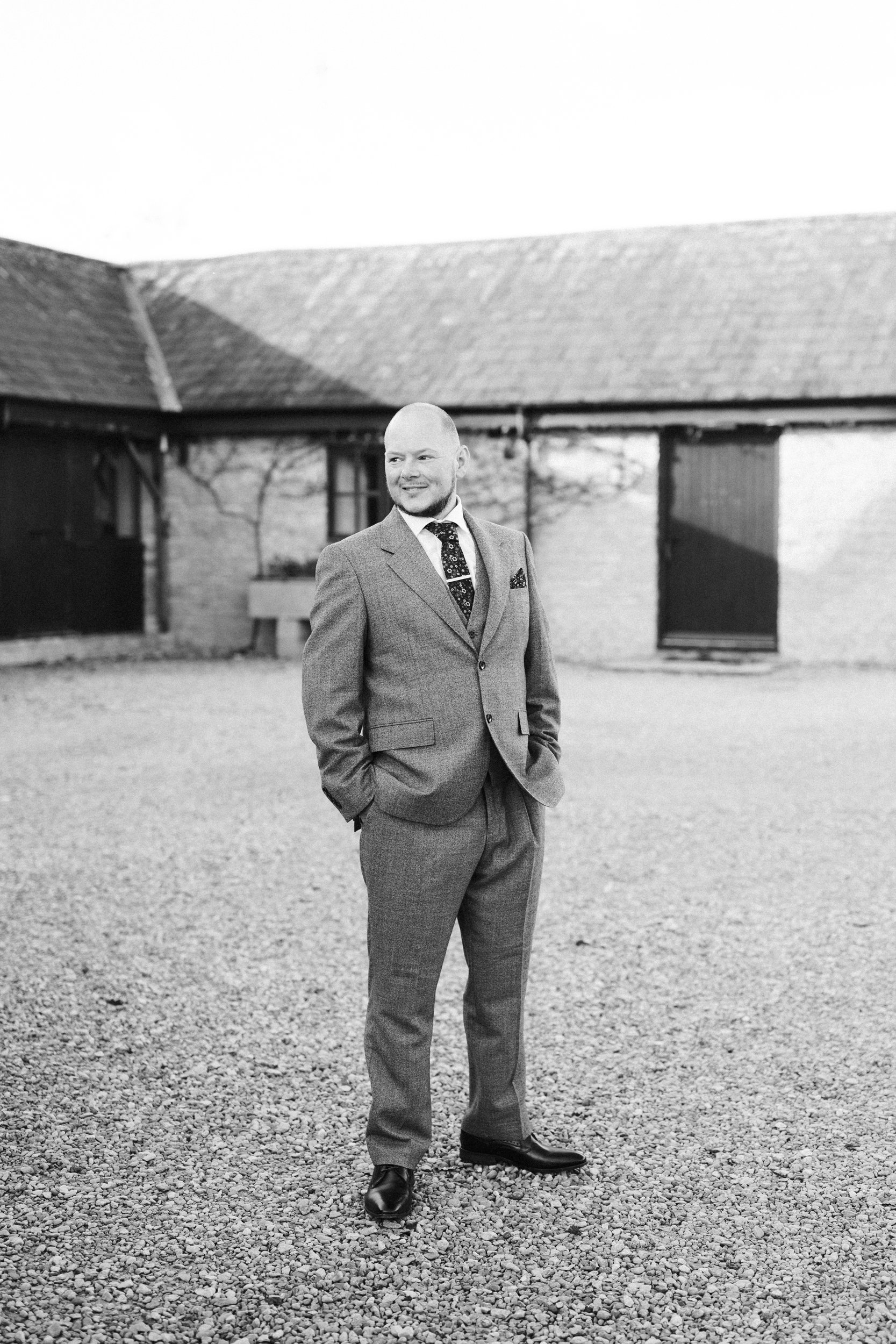 A man in a suit standing outdoors on a gravel surface, with a building in the background.