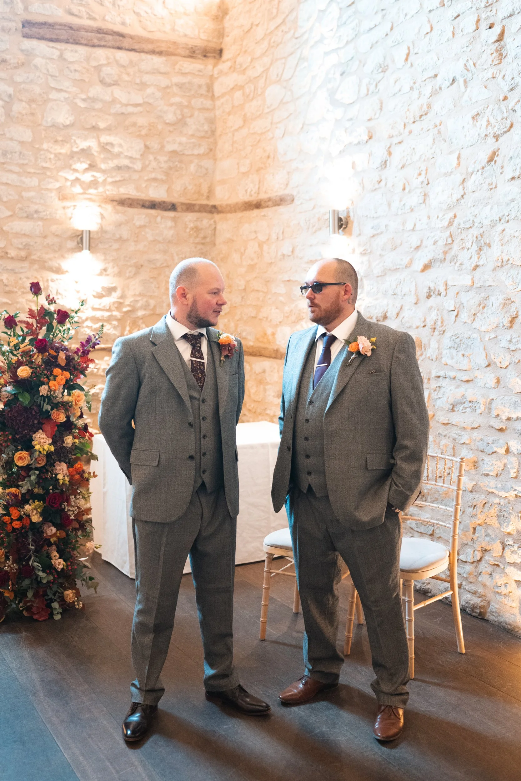 Two men in gray suits talking in a decorated indoor setting with stone walls and a floral arrangement.
