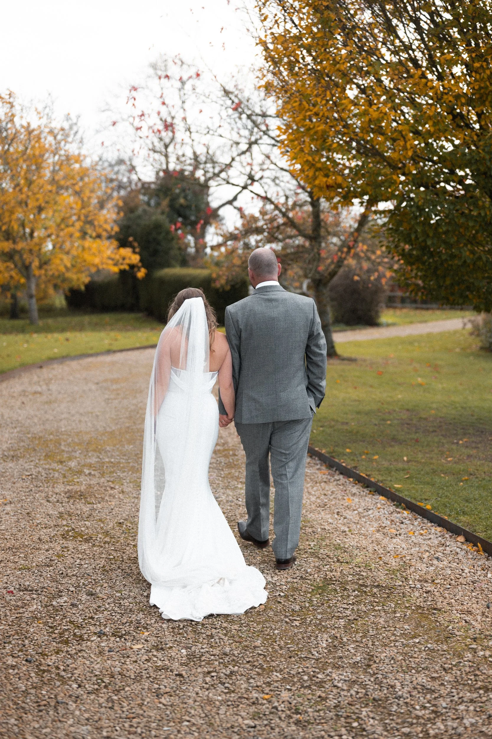 Bride and groom holding hands walking on a gravel path in an outdoor setting during autumn with colorful trees.