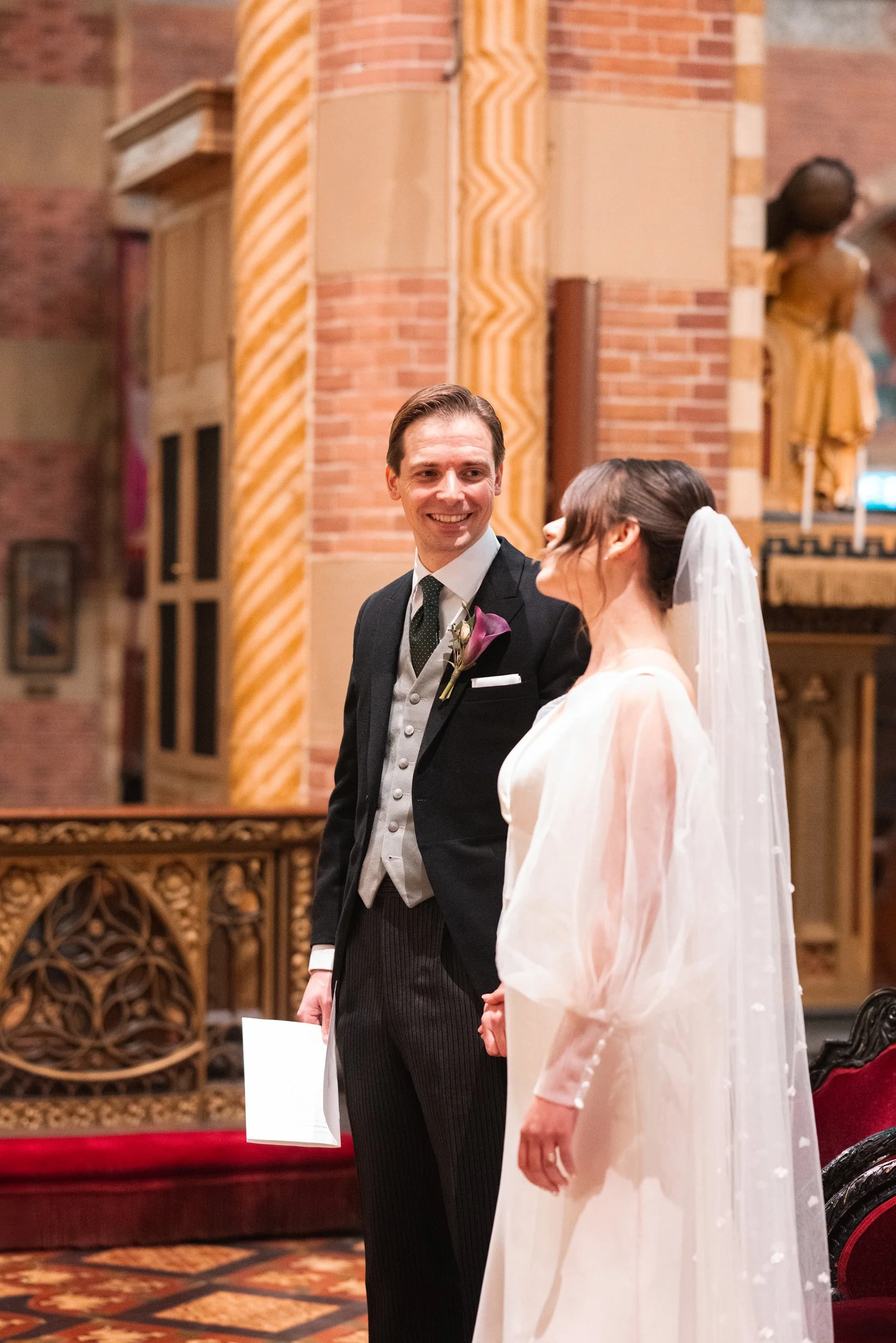 A bride and groom holding hands during their wedding ceremony inside a church, smiling at each other.