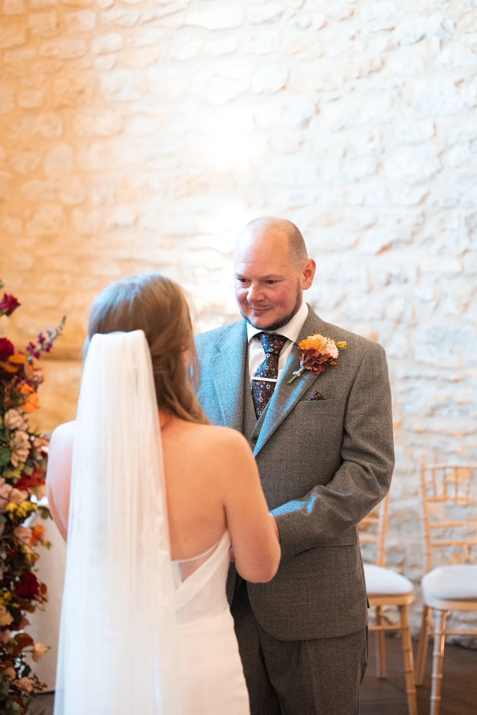 A bride and groom exchanging vows during their wedding ceremony indoors with a brick wall background. The groom is dressed in a gray suit with a floral boutonniere, and the bride in a white dress with a long veil.