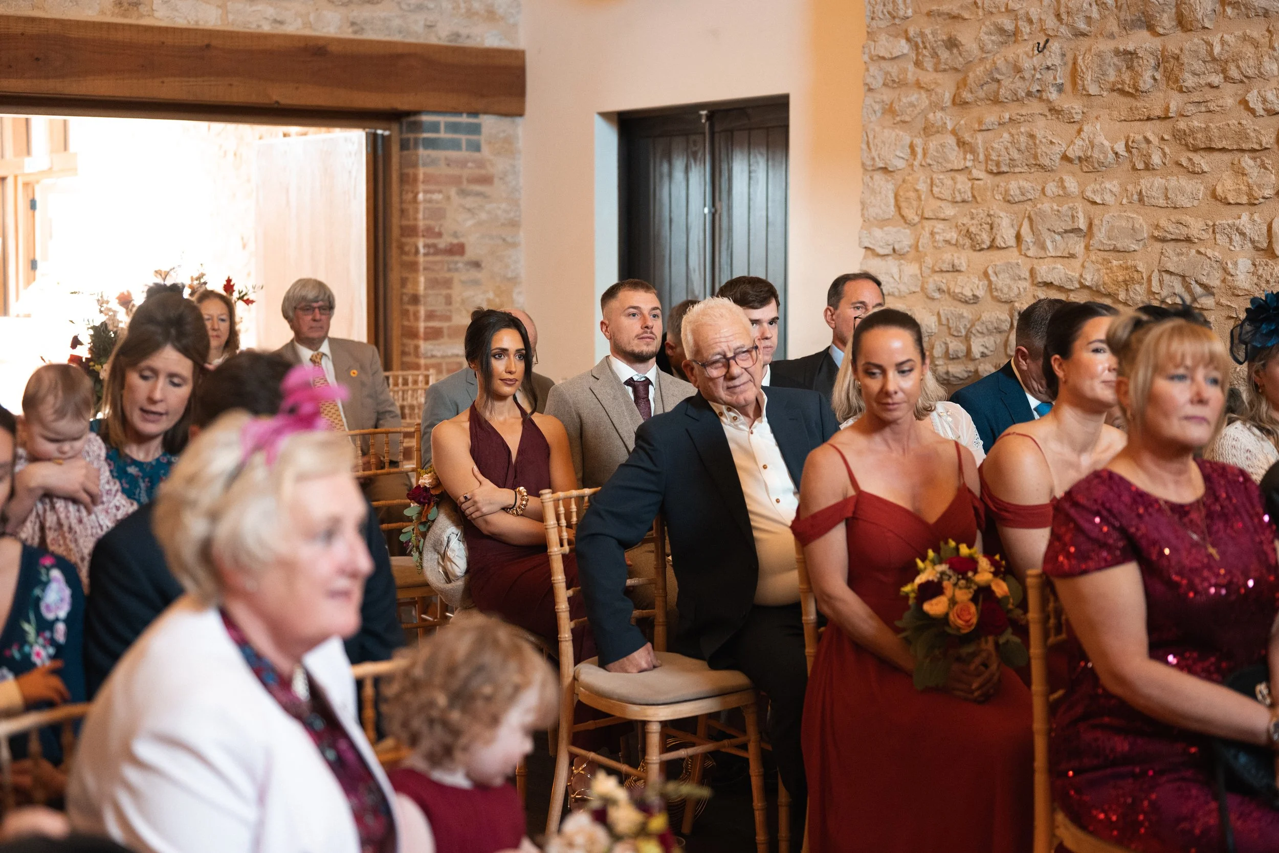 People attending a wedding ceremony sitting on wooden chairs inside a rustic brick and stone building.