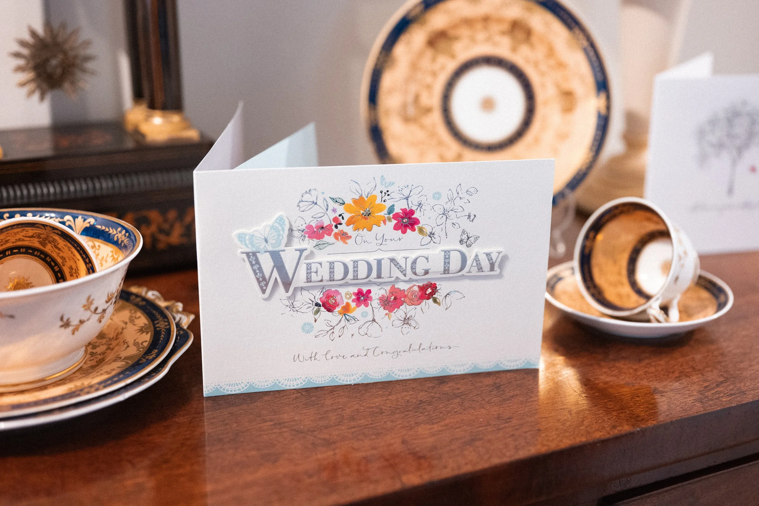 Wedding day greeting card placed on a wooden table, surrounded by decorative fine china cups and plates with ornate patterns, and a large decorative plate or artwork in the background.