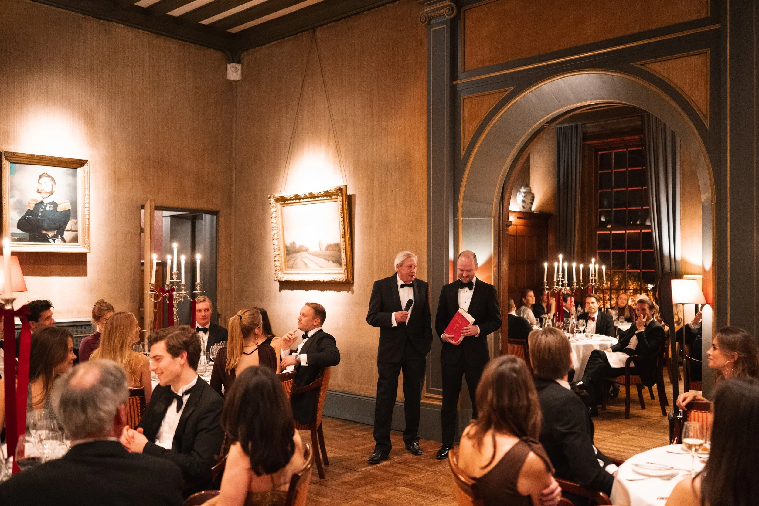 People gathered in a formal dining room for an event, with two men in tuxedos standing and speaking in front of the room, surrounded by seated guests. The room features warm lighting, framed artwork, and elegant decor.