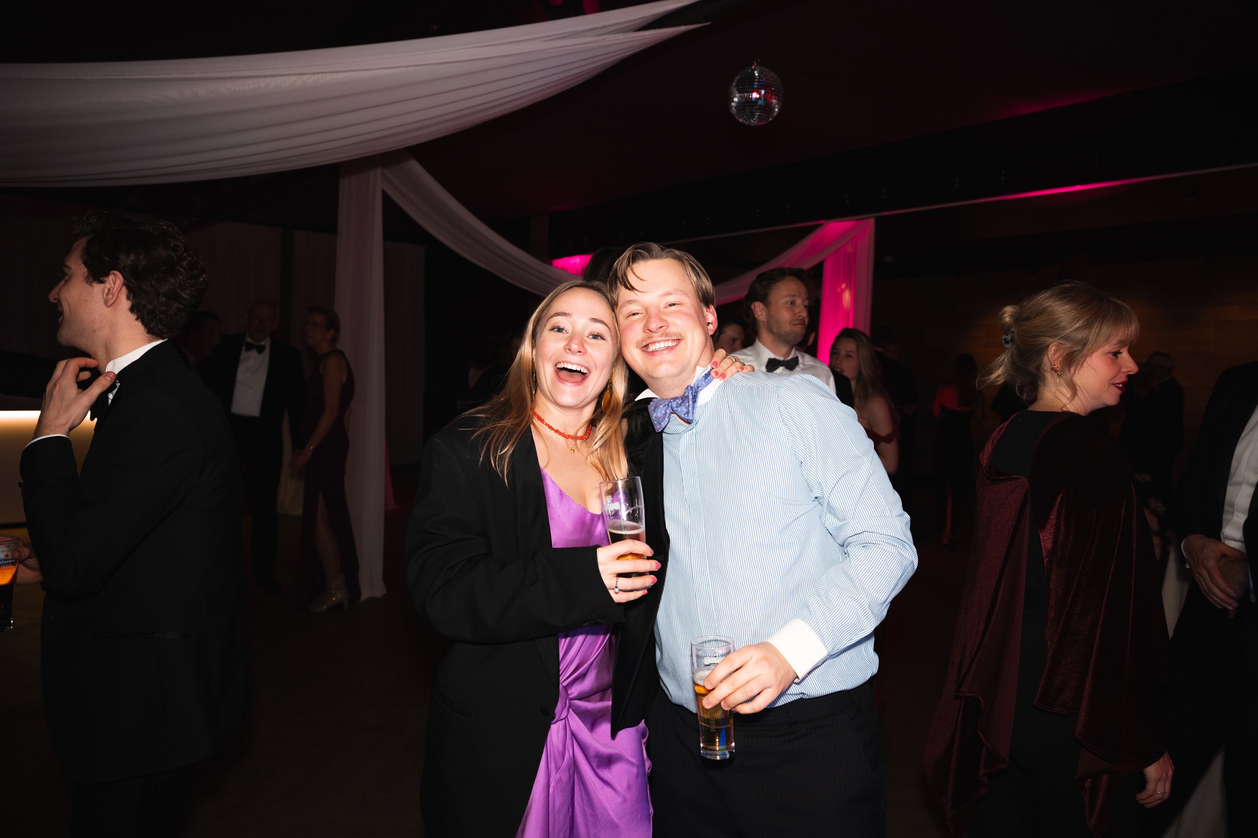 Two young adults, a woman in a purple dress and a man in a light blue shirt with a bow tie, smiling and holding drinks, celebrating at a formal party with other guests in the background.