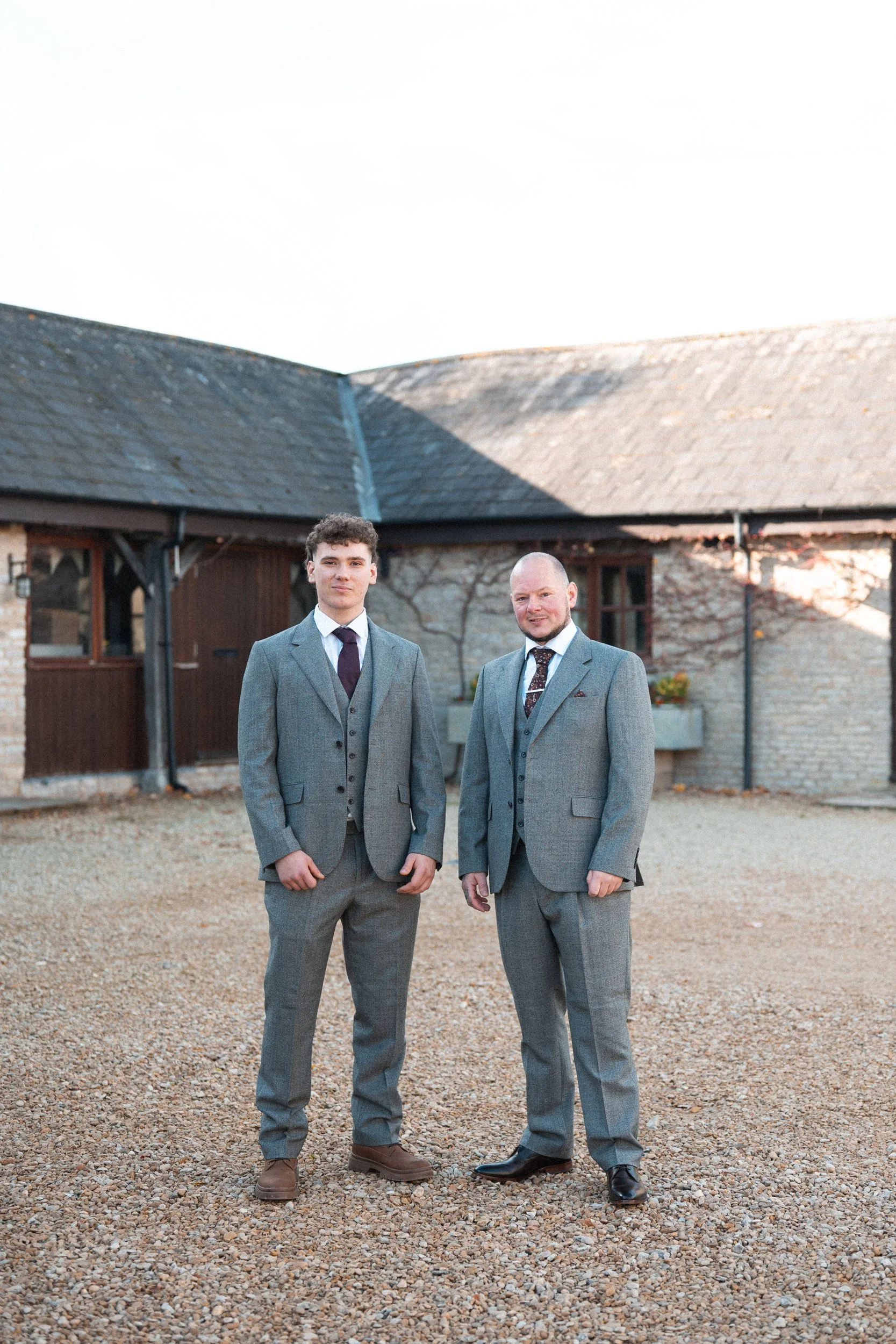 Two men in gray suits standing outside on a gravel surface in front of a rustic building during daylight.