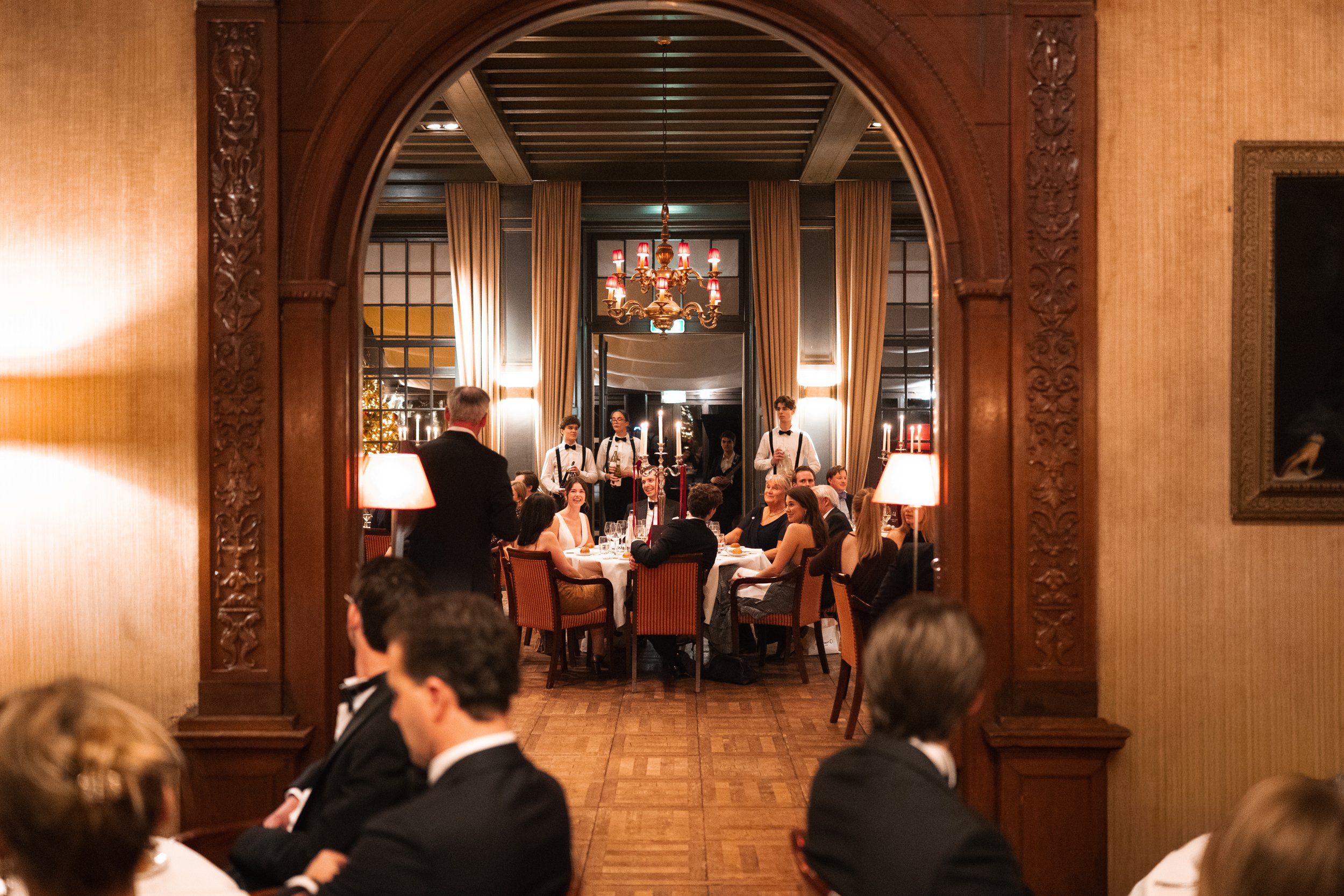 An elegant dining room with a round table full of guests, waitstaff serving, and a decorated Christmas tree visible outside. The scene is viewed through a wooden archway.