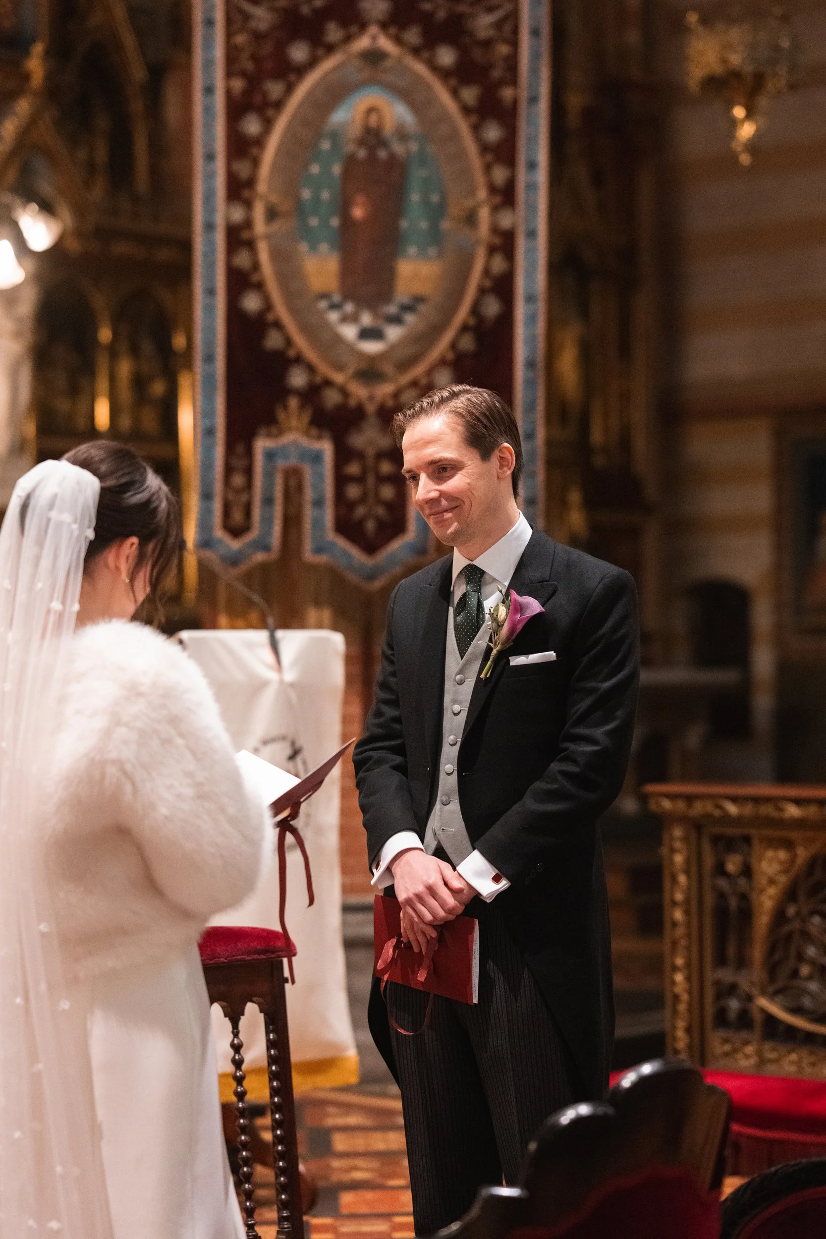 A groom in a tuxedo with a purple boutonniere stands smiling at a bride during a wedding ceremony inside a church with ornate woodwork and religious art in the background.