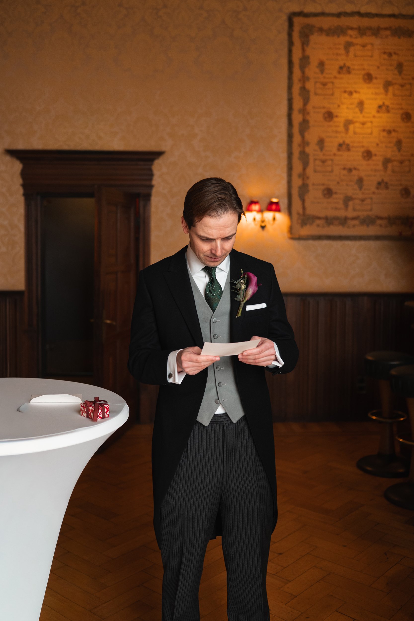 A young man in formal attire, with a flower pinned to his lapel, reading a note at a reception or event in a cozy, dimly-lit room with vintage decor.