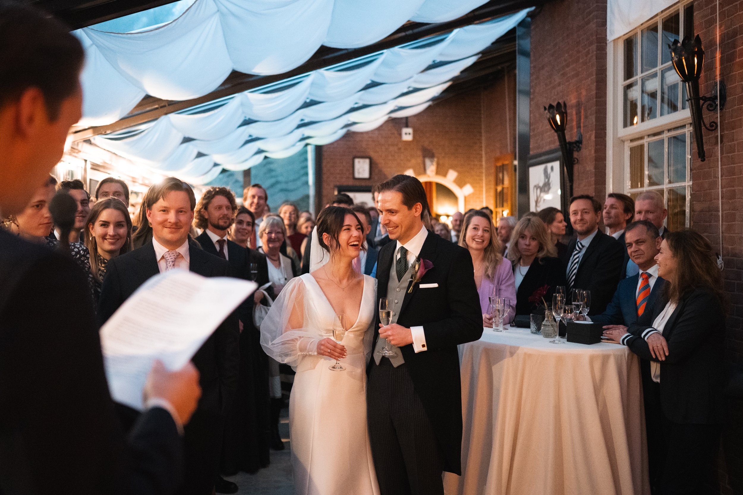 Bride and groom smiling at each other during their wedding reception, surrounded by guests in a decorated indoor venue.