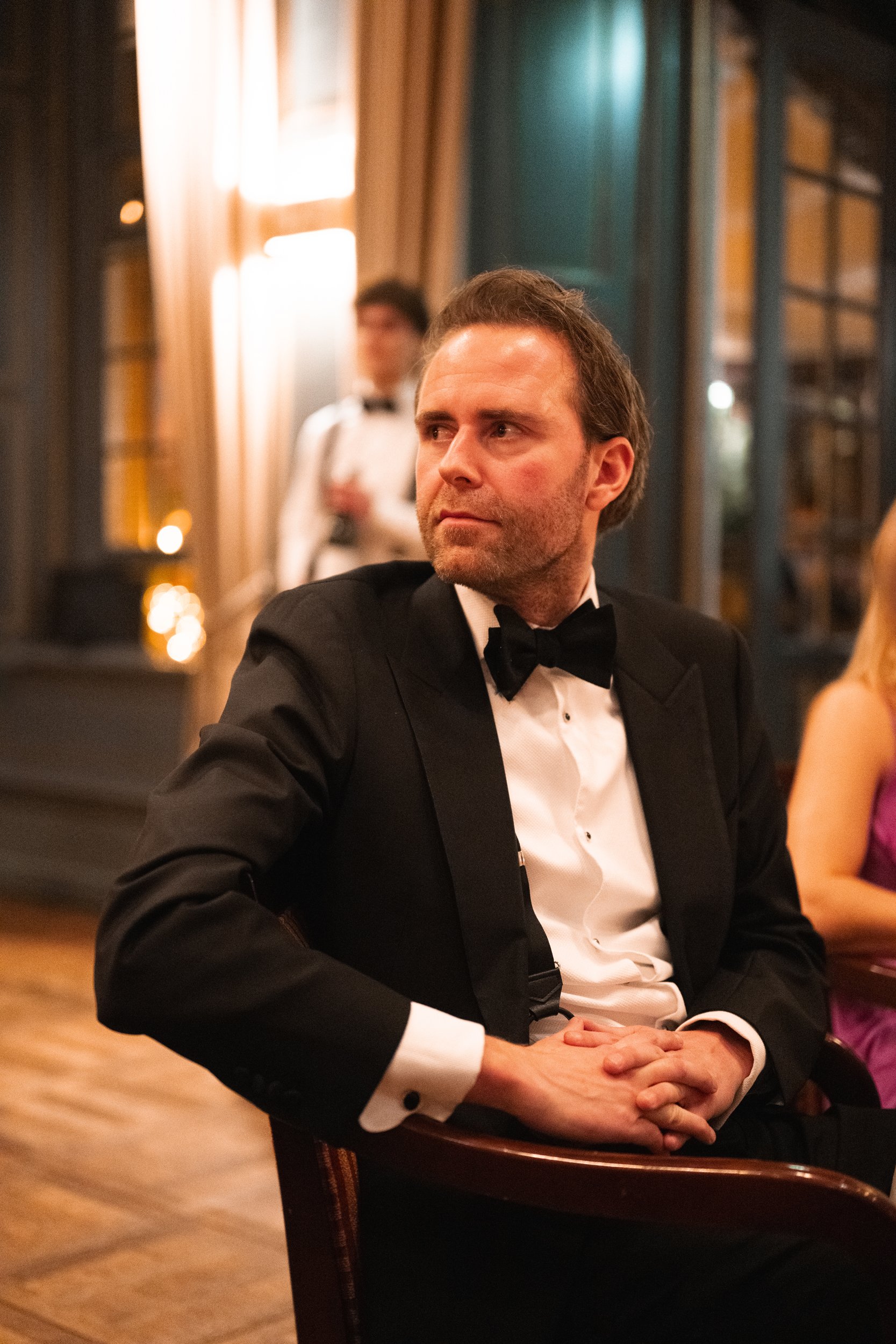 A man in a black tuxedo with a bow tie sitting at a formal event, with a woman in purple beside him and a waiter in the background.