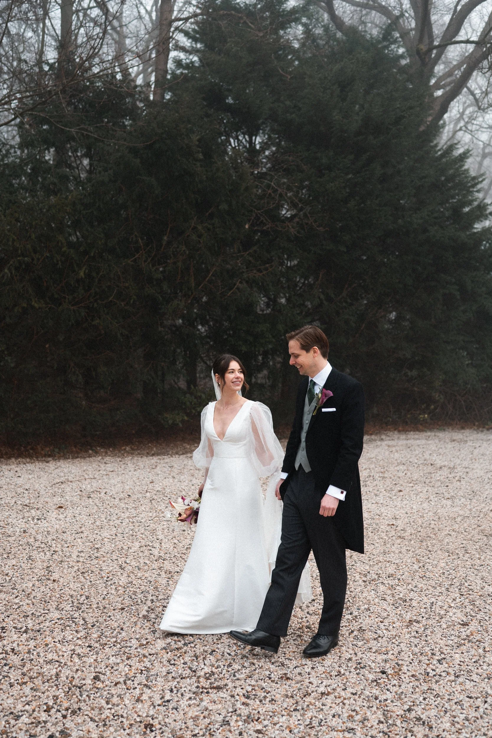 A bride and groom walking together outdoors, holding hands, with the bride holding a bouquet, surrounded by trees and a foggy atmosphere.