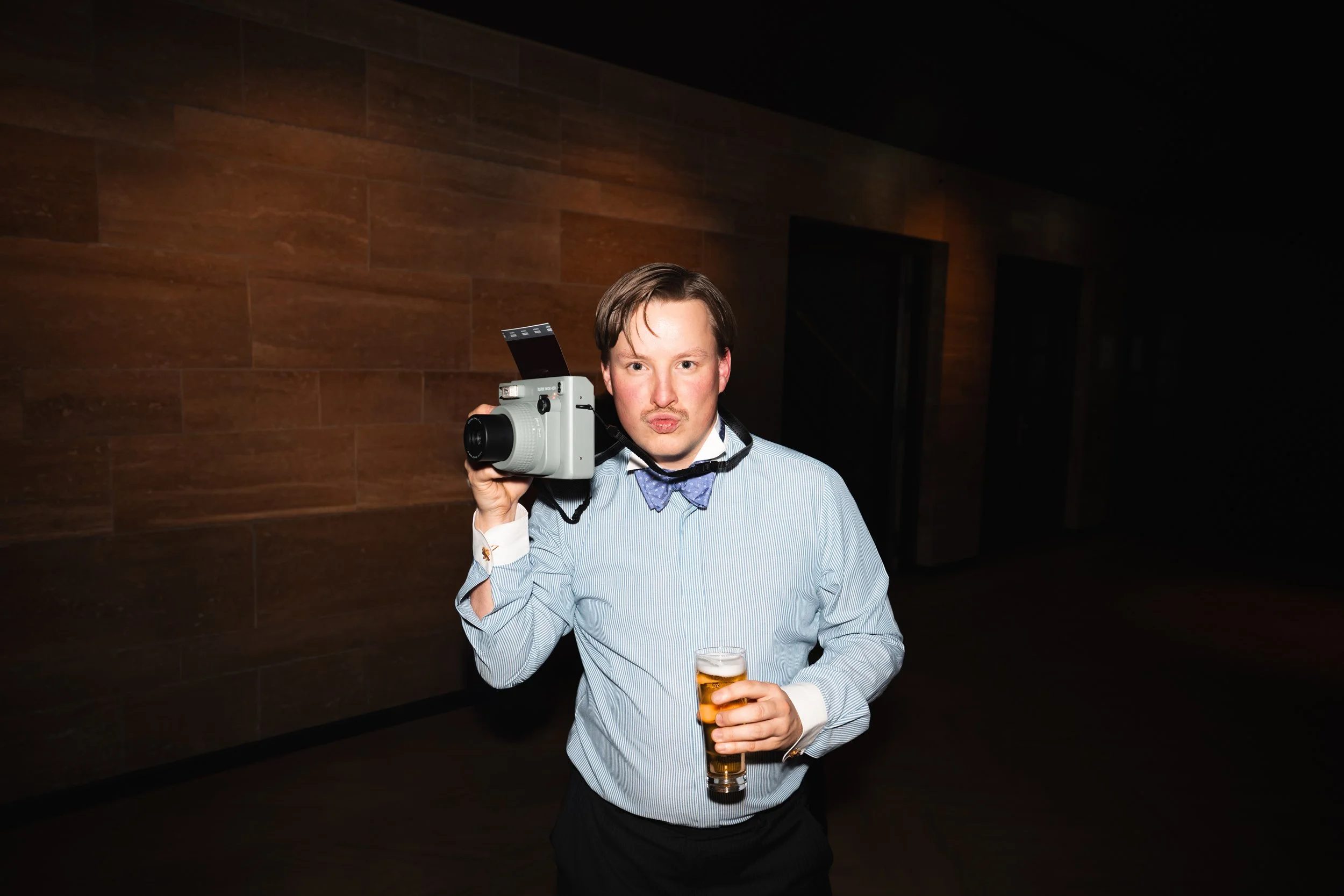 A man in a dress shirt with a bow tie holding a camera over his shoulder and a beer in his other hand, standing in a dimly lit indoor space.