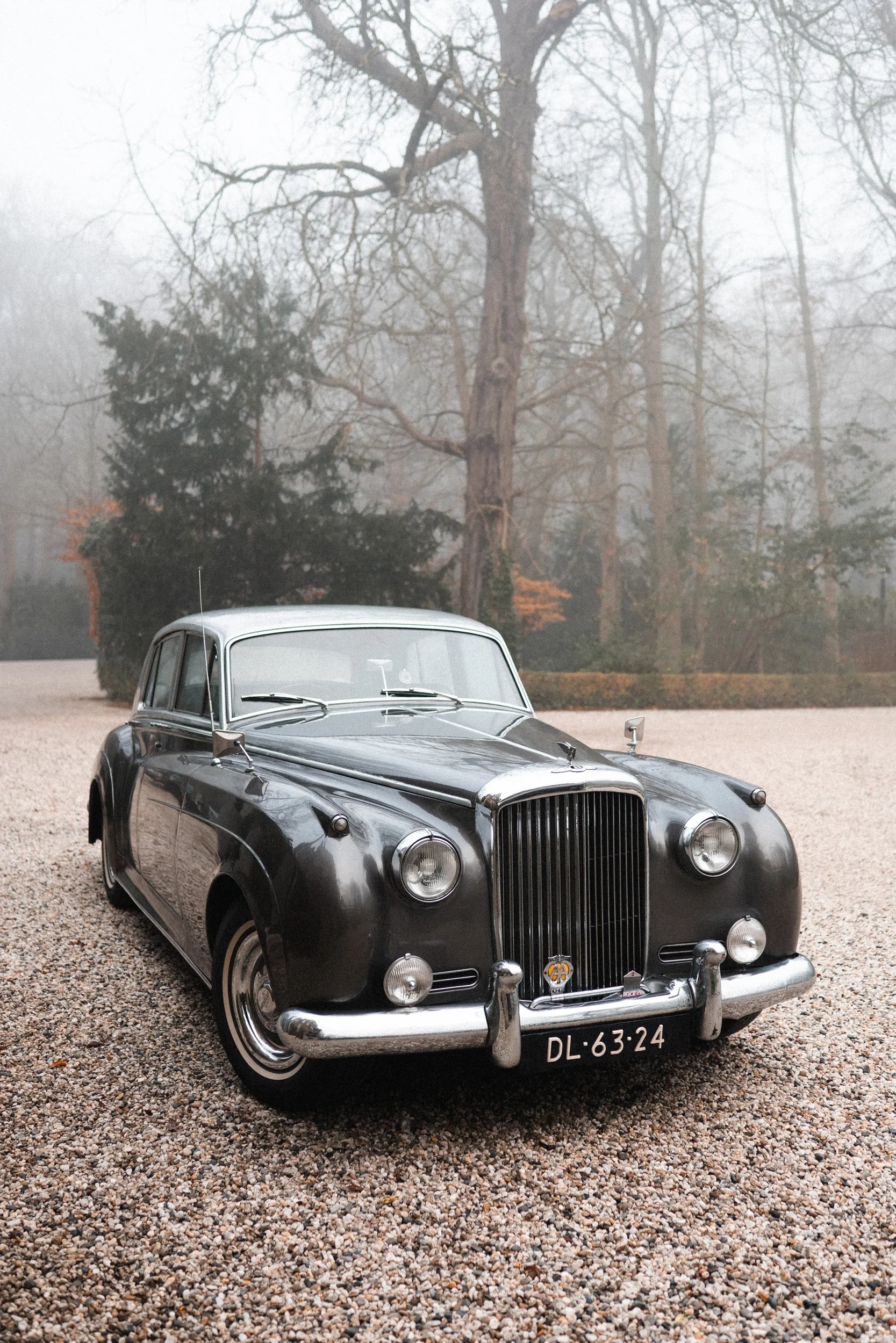 A vintage black car parked on gravel with trees and fog in the background.