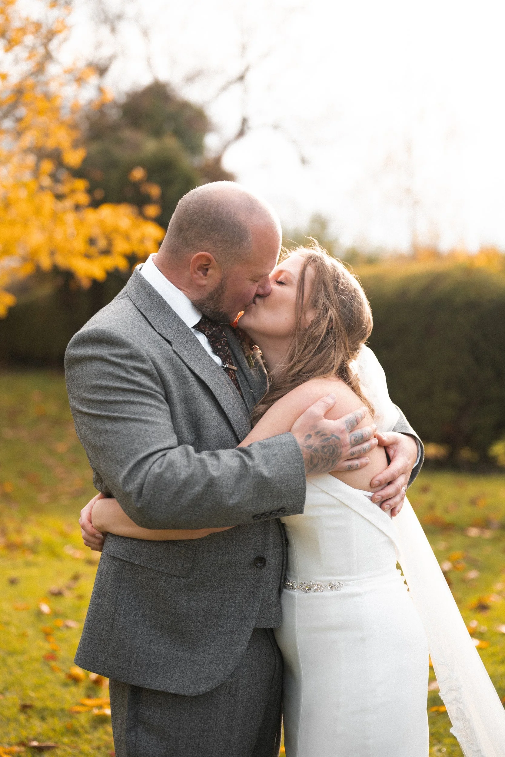 A newlywed couple sharing a kiss outdoors in fall, with the groom wearing a gray suit and the bride in a white wedding dress, surrounded by autumn leaves.
