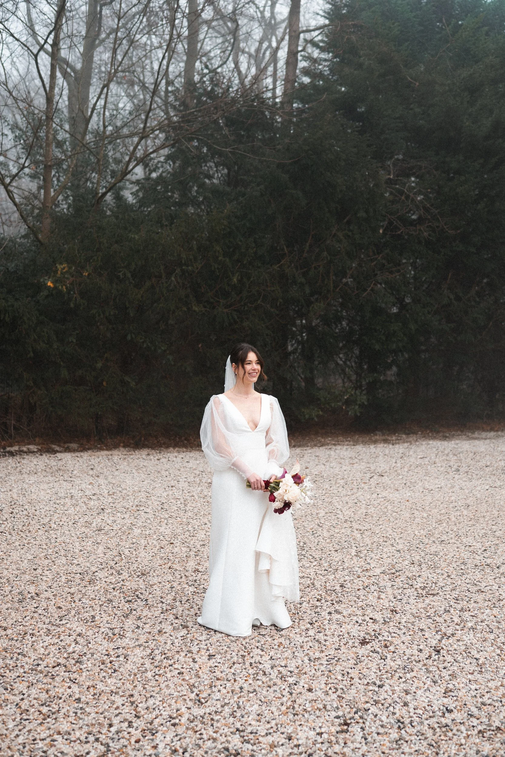 Bride in a white wedding dress holding a bouquet, standing on a gravel surface outdoors with trees and fog in the background.