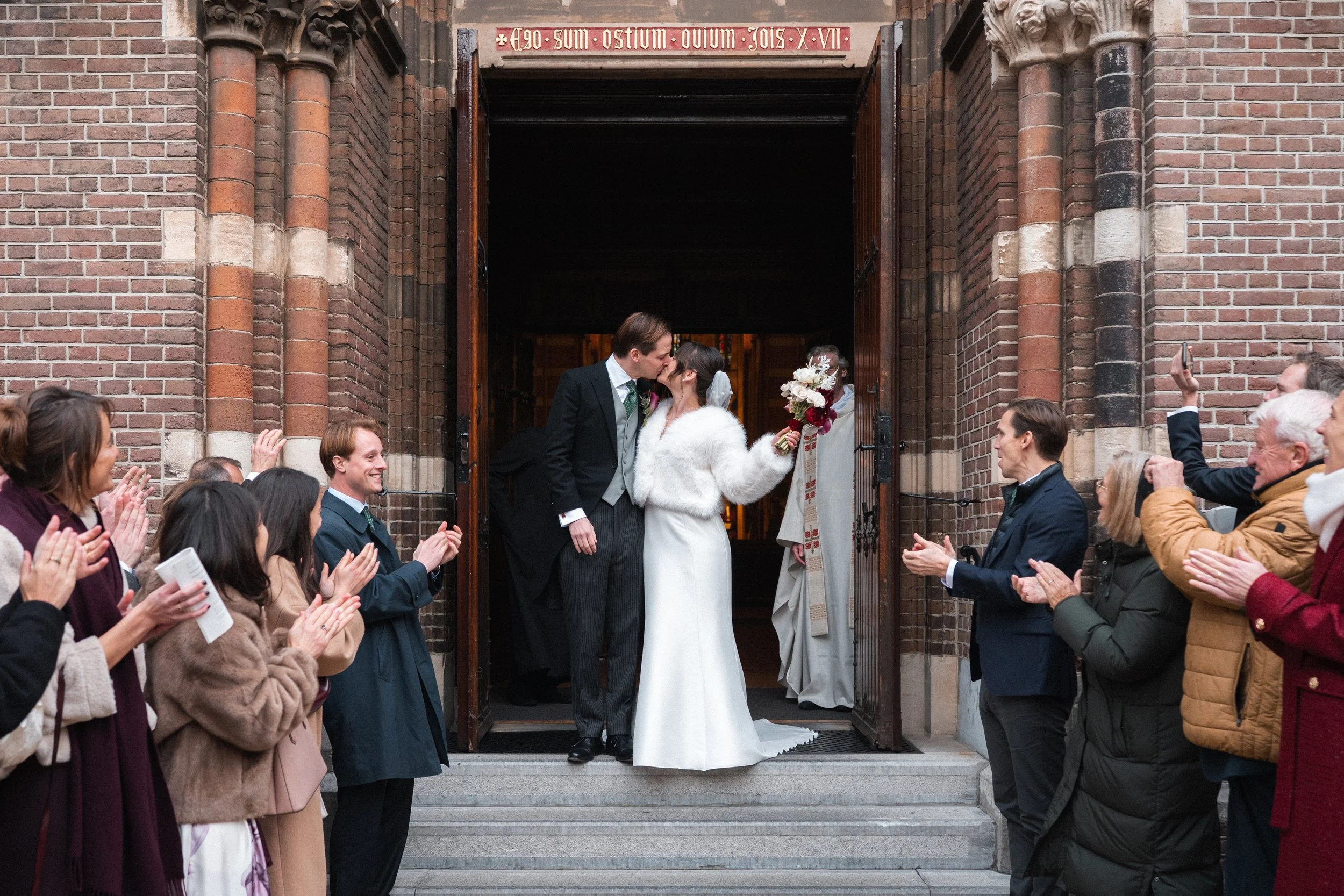A bride and groom share a kiss on the church steps, surrounded by clapping friends and family during their wedding celebration.