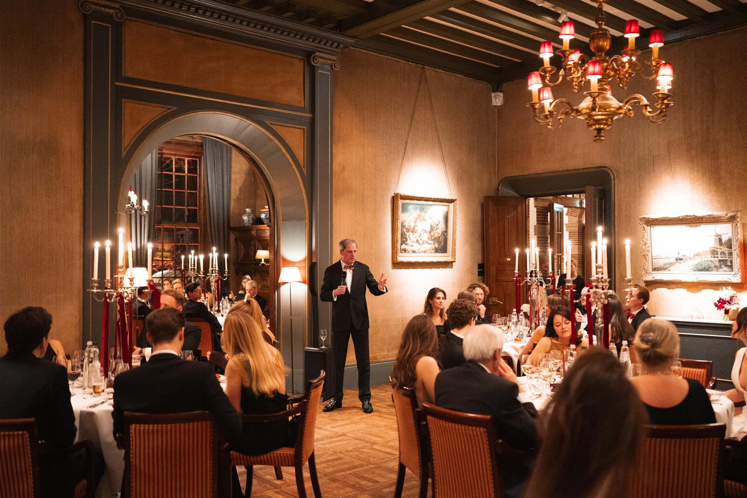 An elegant dinner event in a luxurious room with high ceilings, chandeliers, and warm lighting, featuring guests seated at round tables and a man in a tuxedo giving a speech.