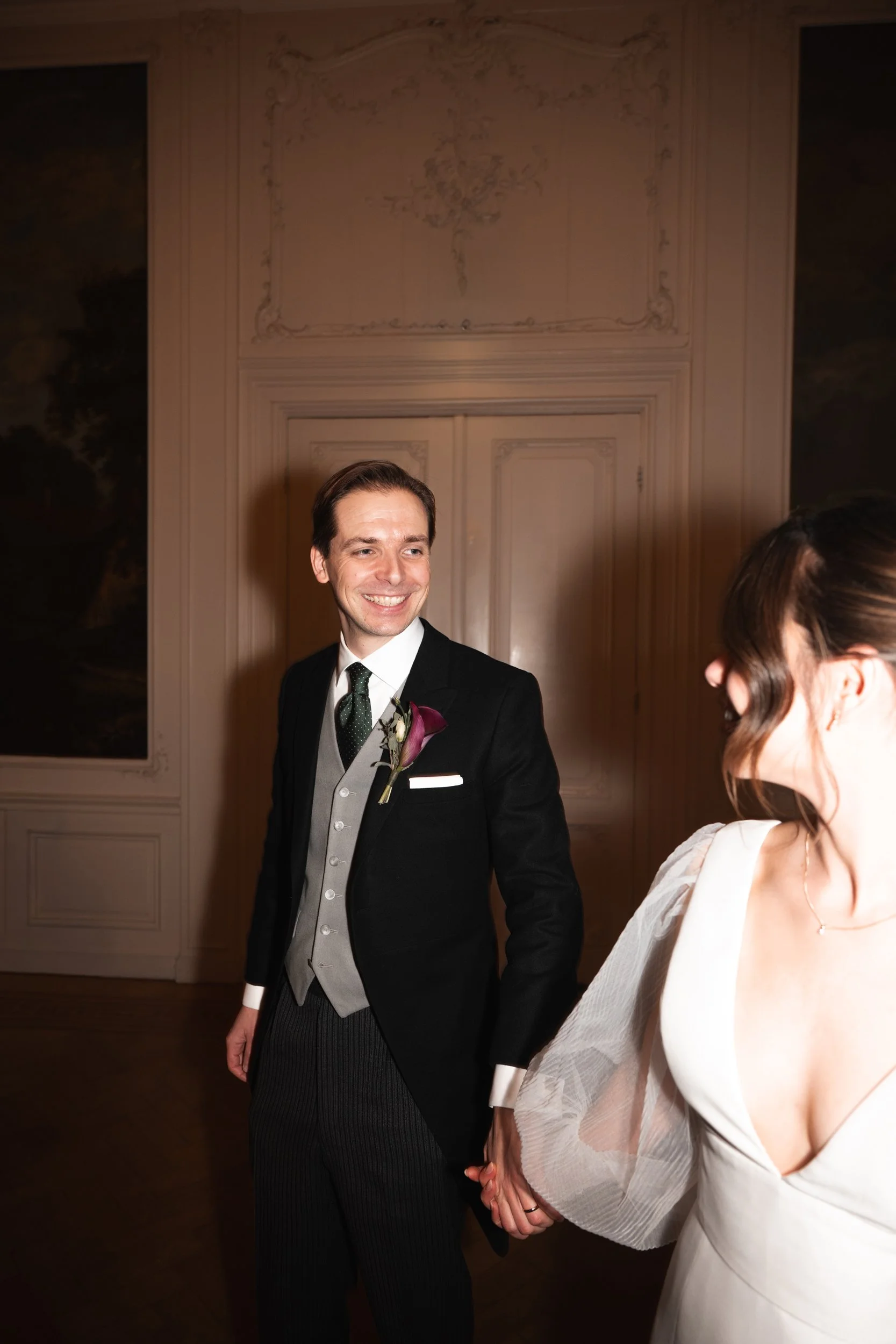 Smiling groom in a black tuxedo holding hands with bride in a white dress, in an elegant room with ornate wall details.