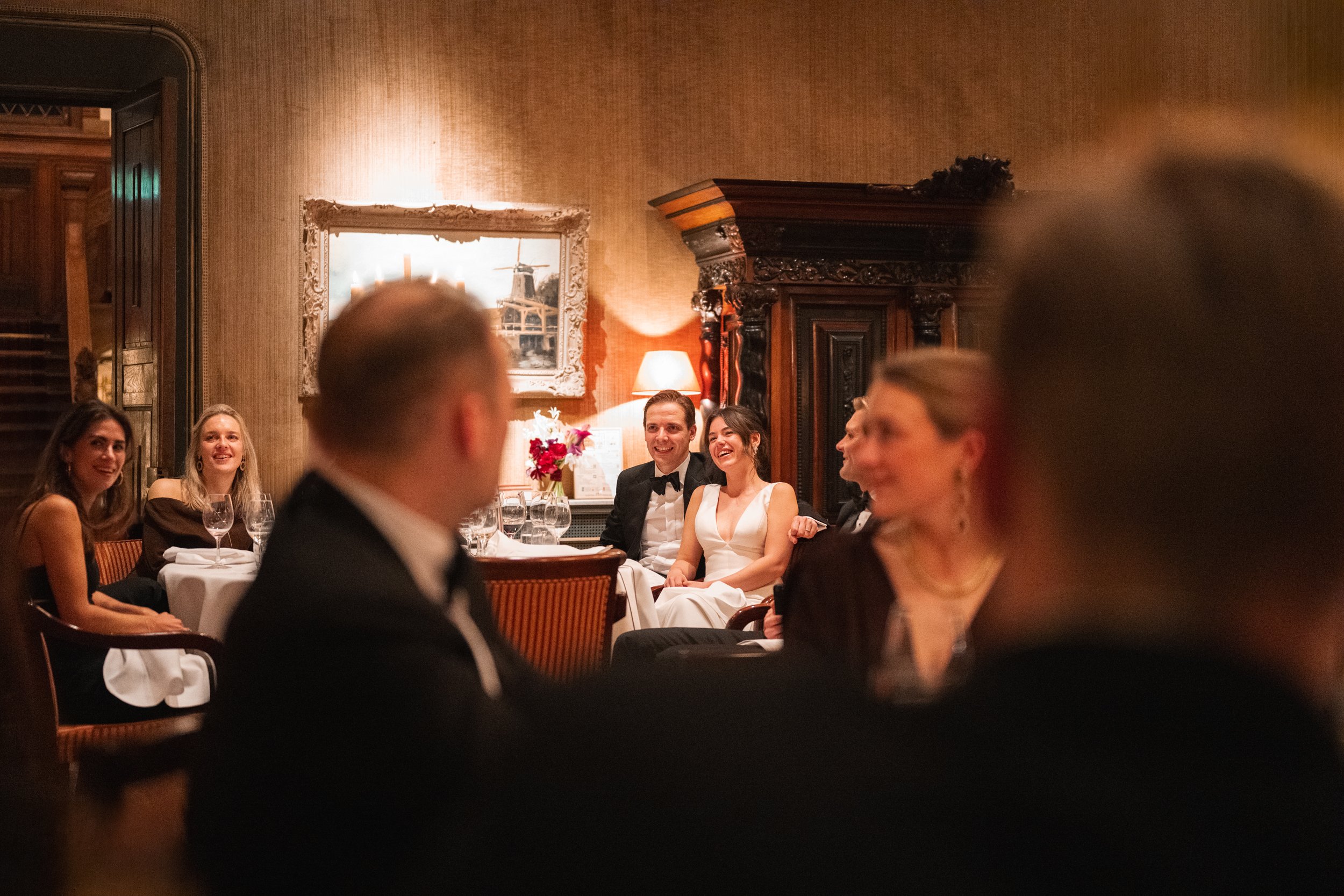 People sitting at a round dinner table in an elegant, warmly lit room, smiling and engaged in conversation.