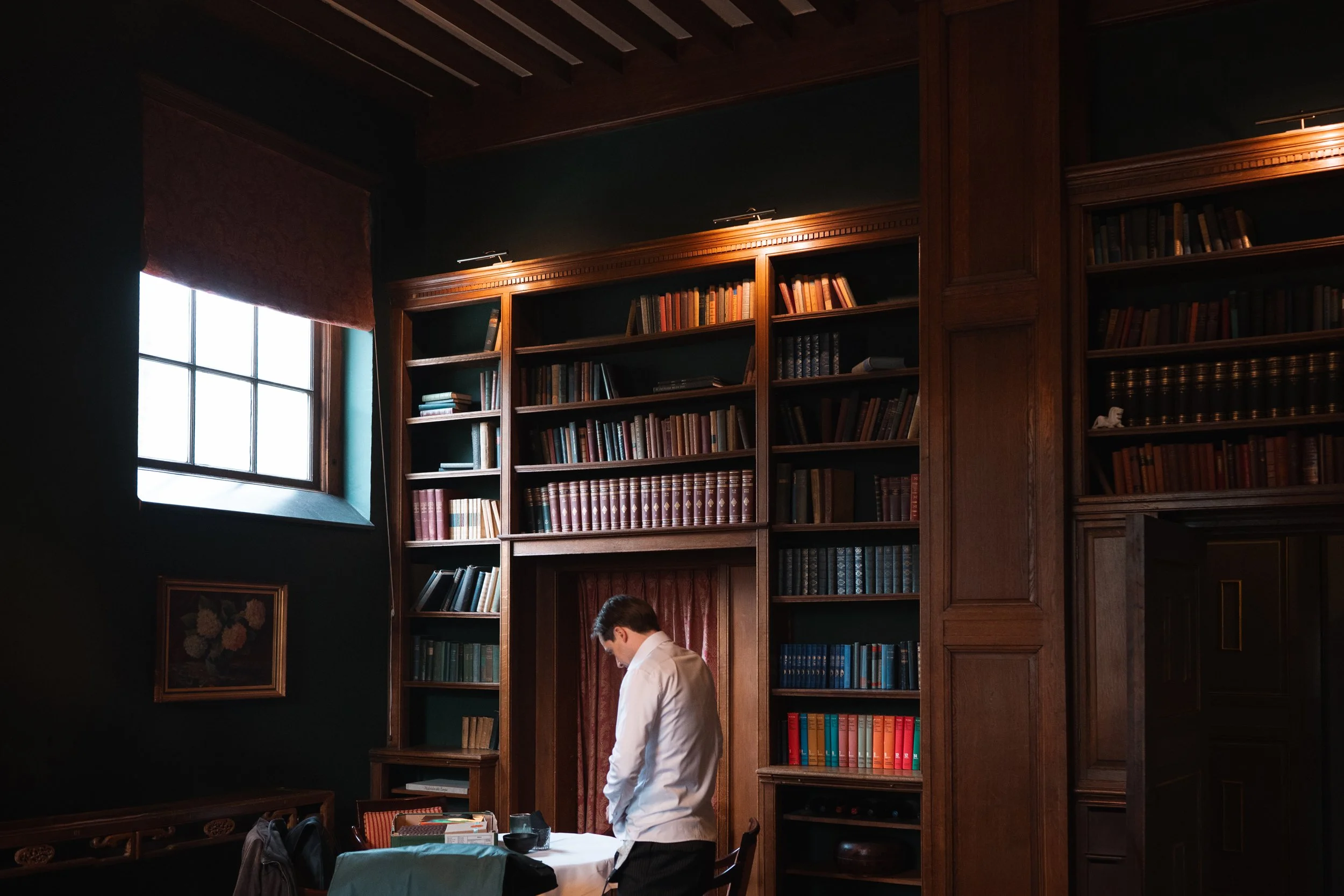 A man in a white shirt standing in a dimly lit, wood-paneled library with tall bookshelves filled with books, near a window with a reddish curtain.
