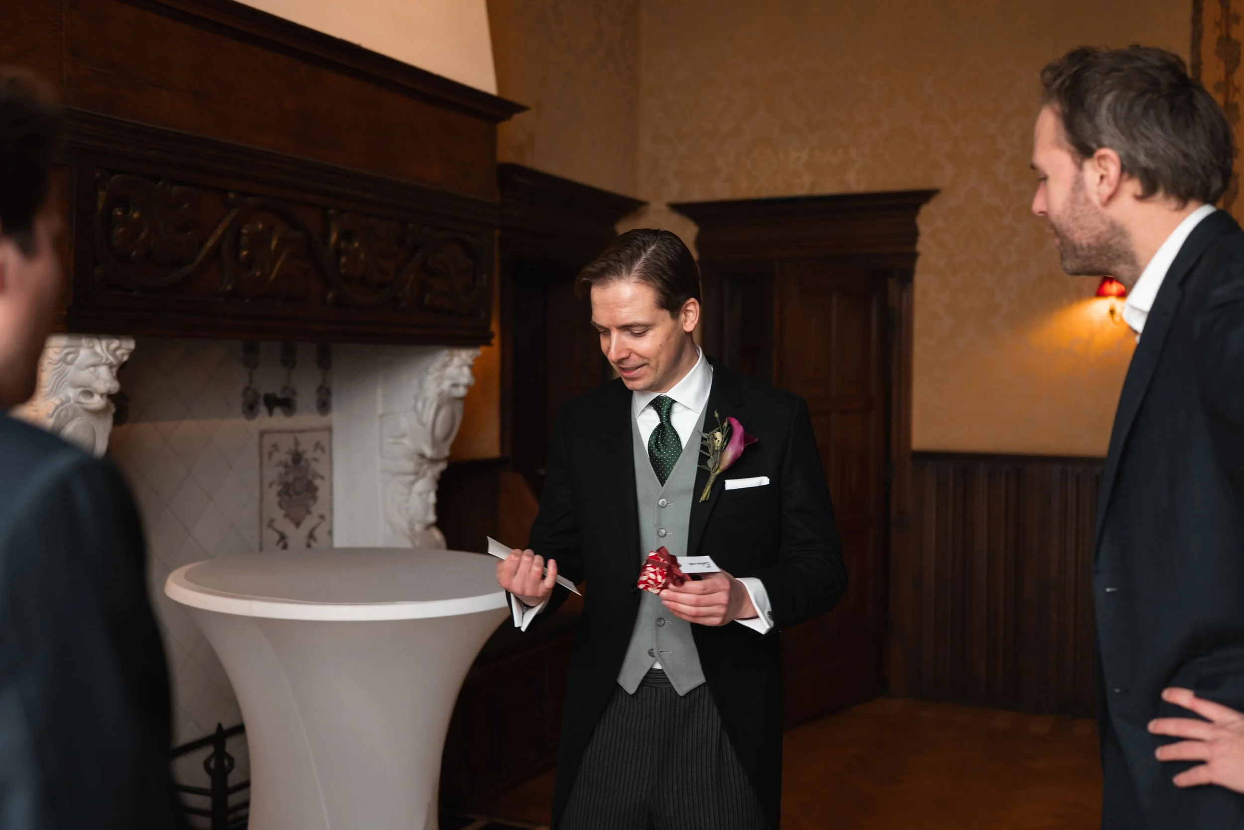 A man dressed in a black suit with a purple calla lily boutonniere is reading from a card while holding a red ribboned gift at a formal indoor setting. Two other men in suits are observing him.