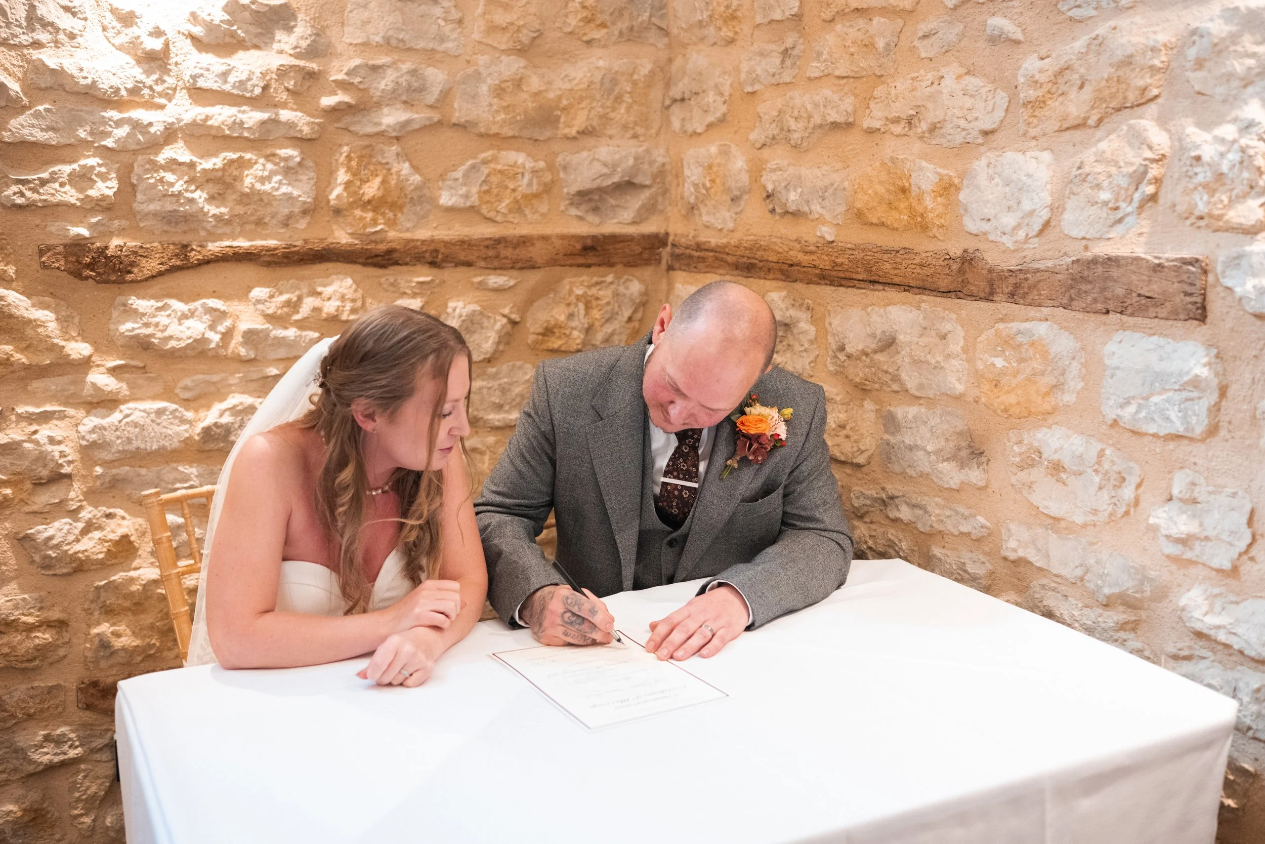 A bride and a groom signing a marriage certificate at a table in a rustic stone-walled room.