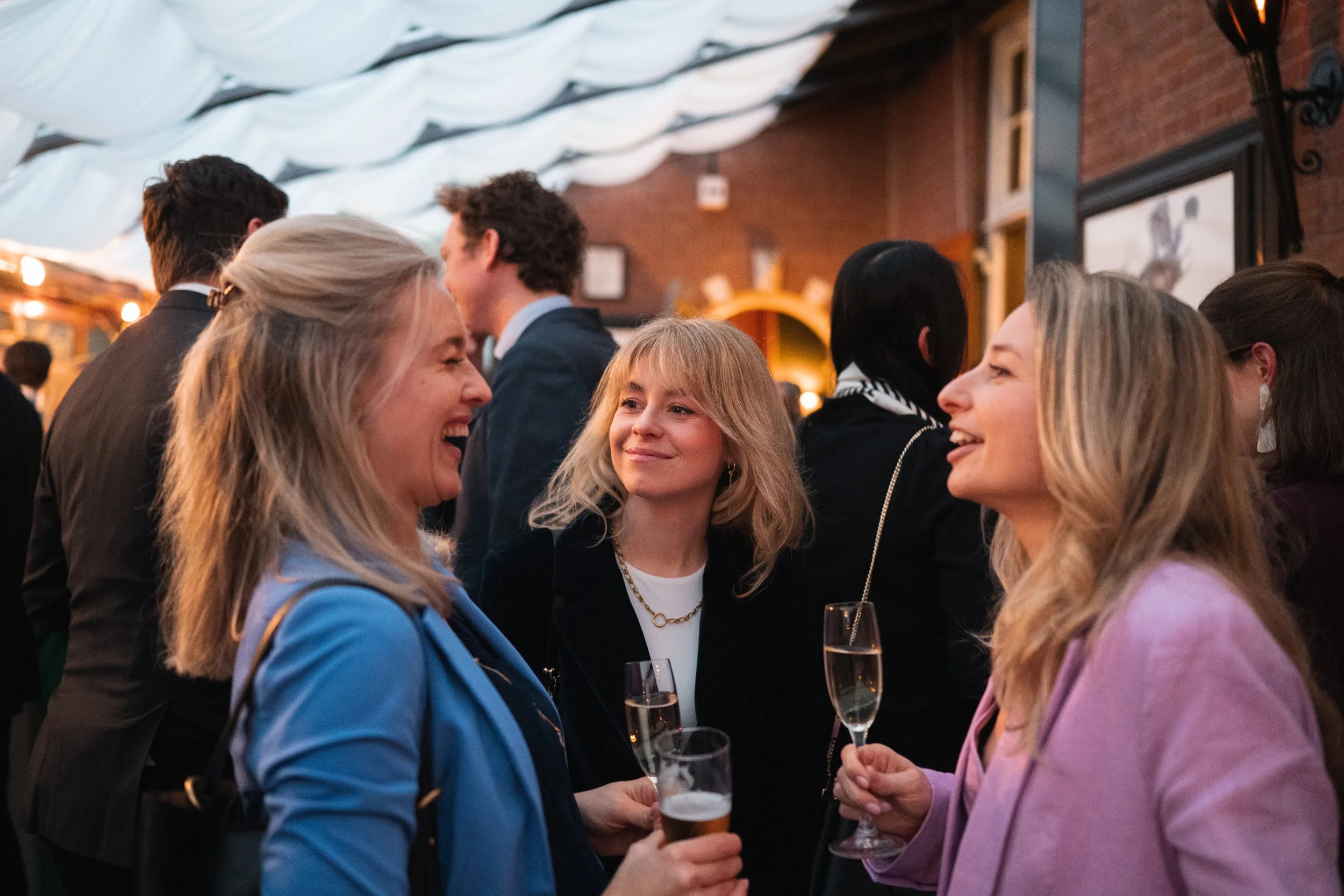 Three women laughing and talking at a social event, holding drinks, with a crowd of people in the background.