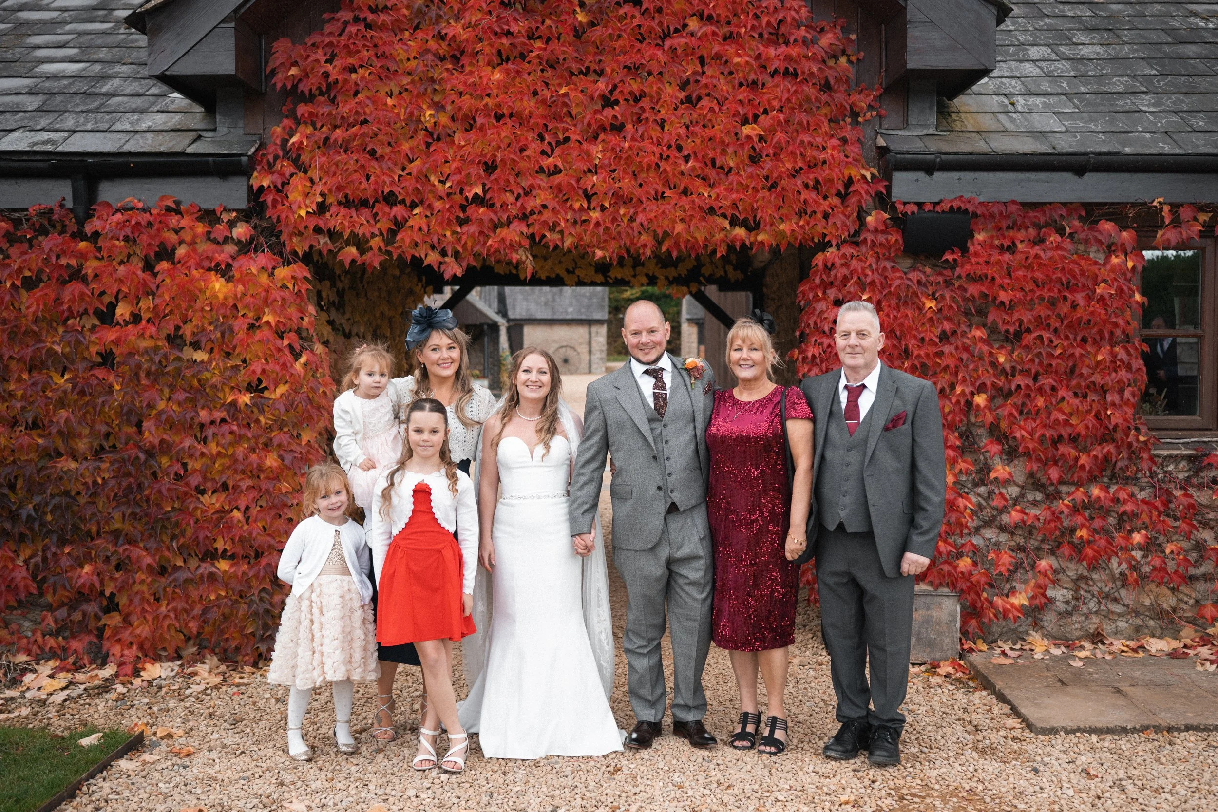 A group of nine people, including a bride in a white wedding gown and a groom in a gray suit, standing outdoors in front of a building covered in red and orange autumn leaves. They are smiling and holding hands.
