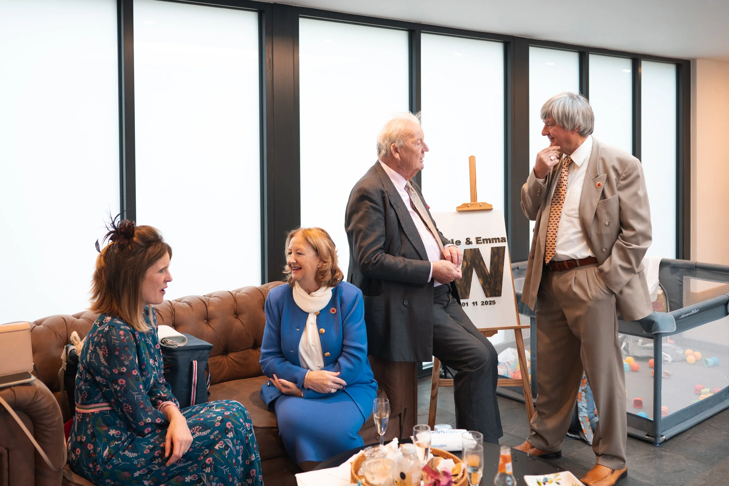 Four people, three women and one man, are engaged in a conversation in a social setting, with drinks and snacks on a table in front of them. The background shows large windows and a guitar in a stand.