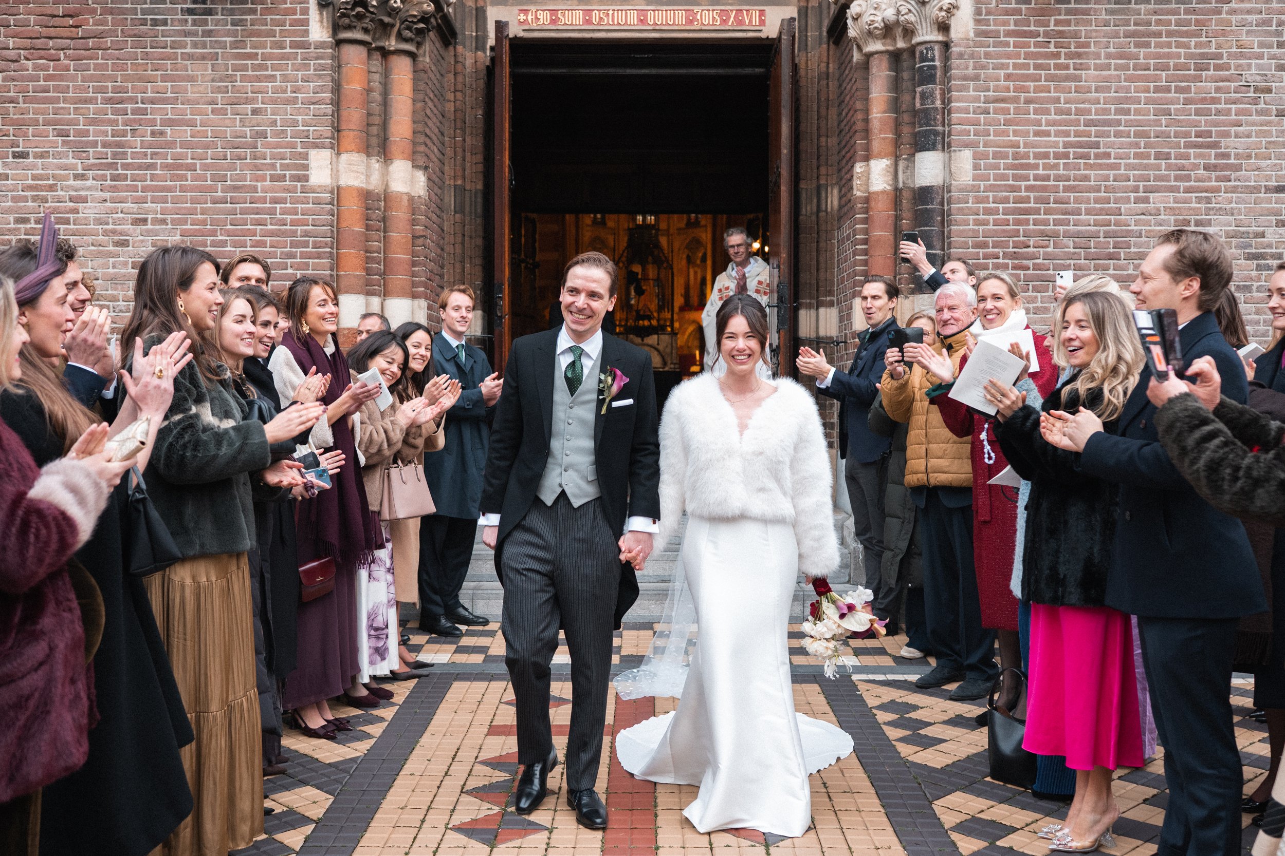 A newly married couple walking out of a church, holding hands, surrounded by cheering friends and family, with some taking photos.