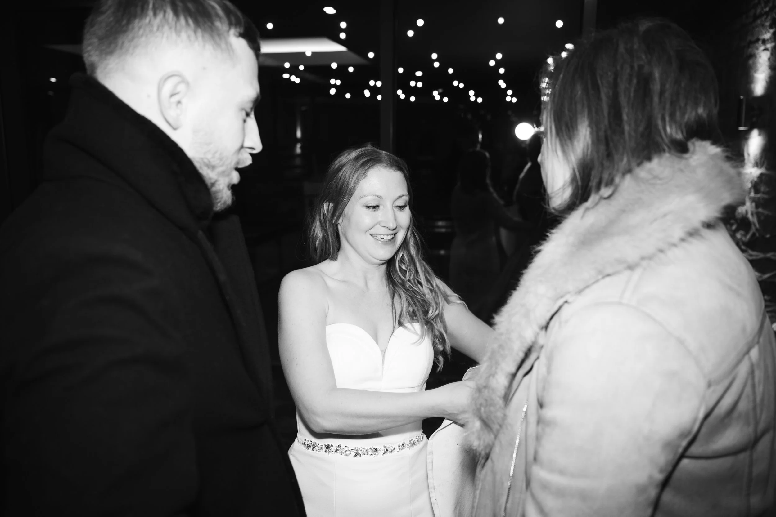Three people at an event, with one woman in a white strapless dress smiling and holding hands with a person in a fur-collared coat, while another man in a dark coat watches.