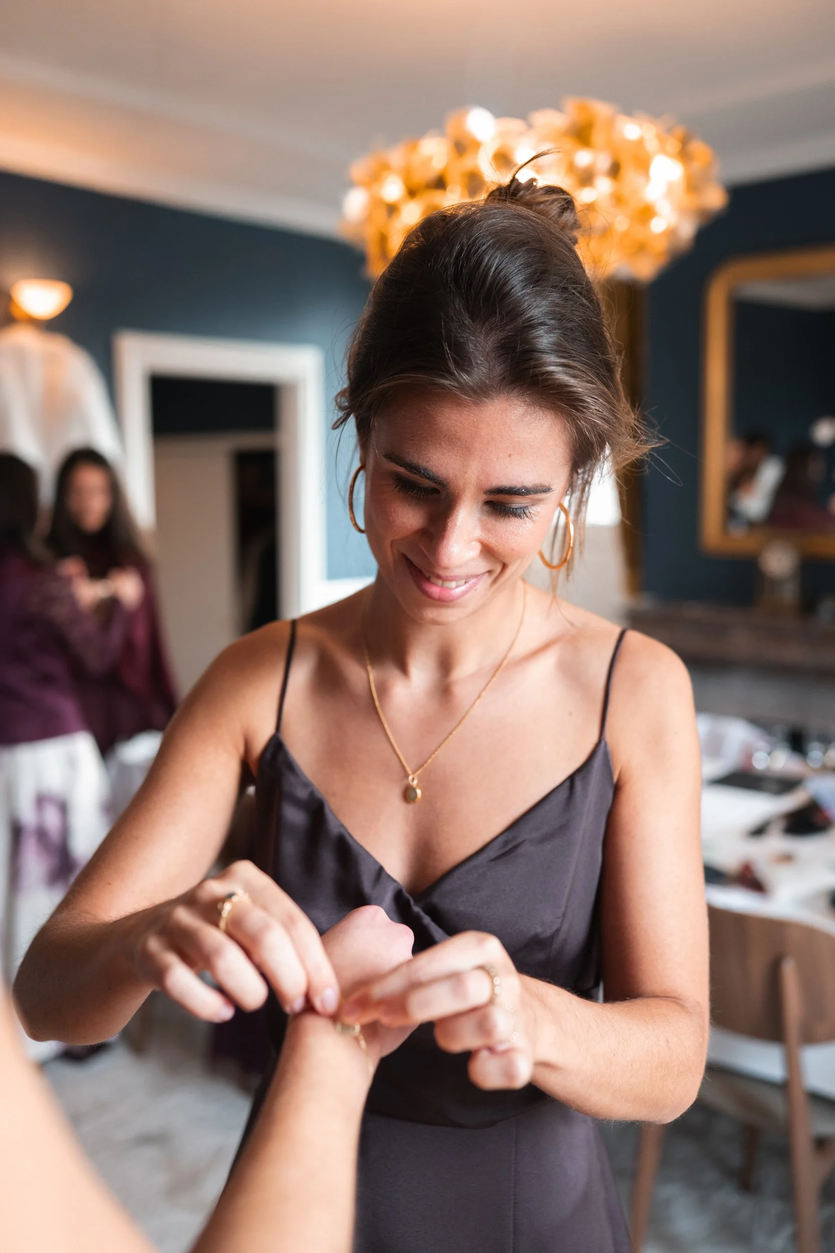 A woman in a black satin dress with gold jewelry, smiling as she places a ring on another person's finger inside a decorated indoor setting.