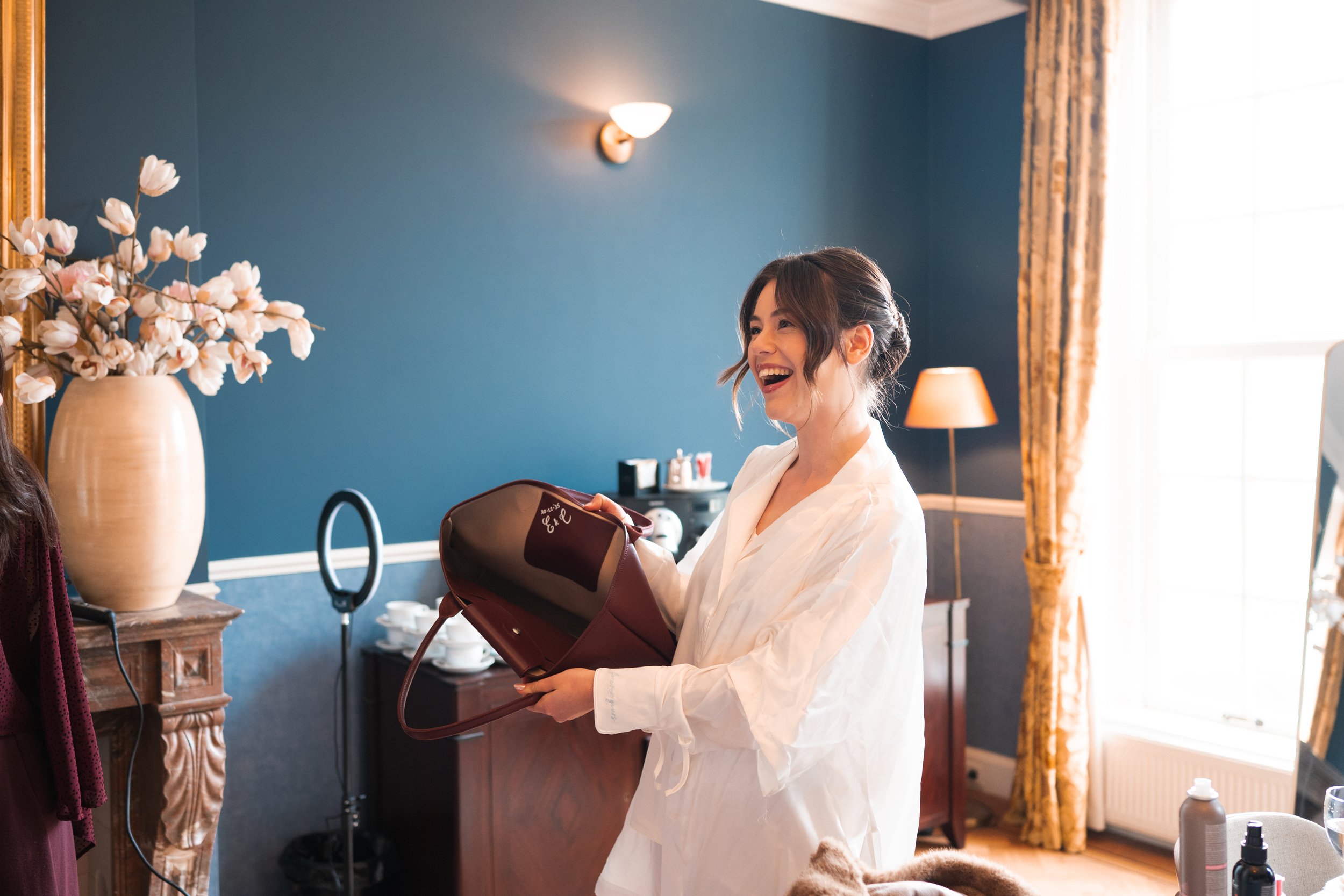 A woman with dark hair in a bun, wearing a white shirt, laughing and holding a maroon purse in a blue-walled room with yellow curtains and a large vase with white flowers.