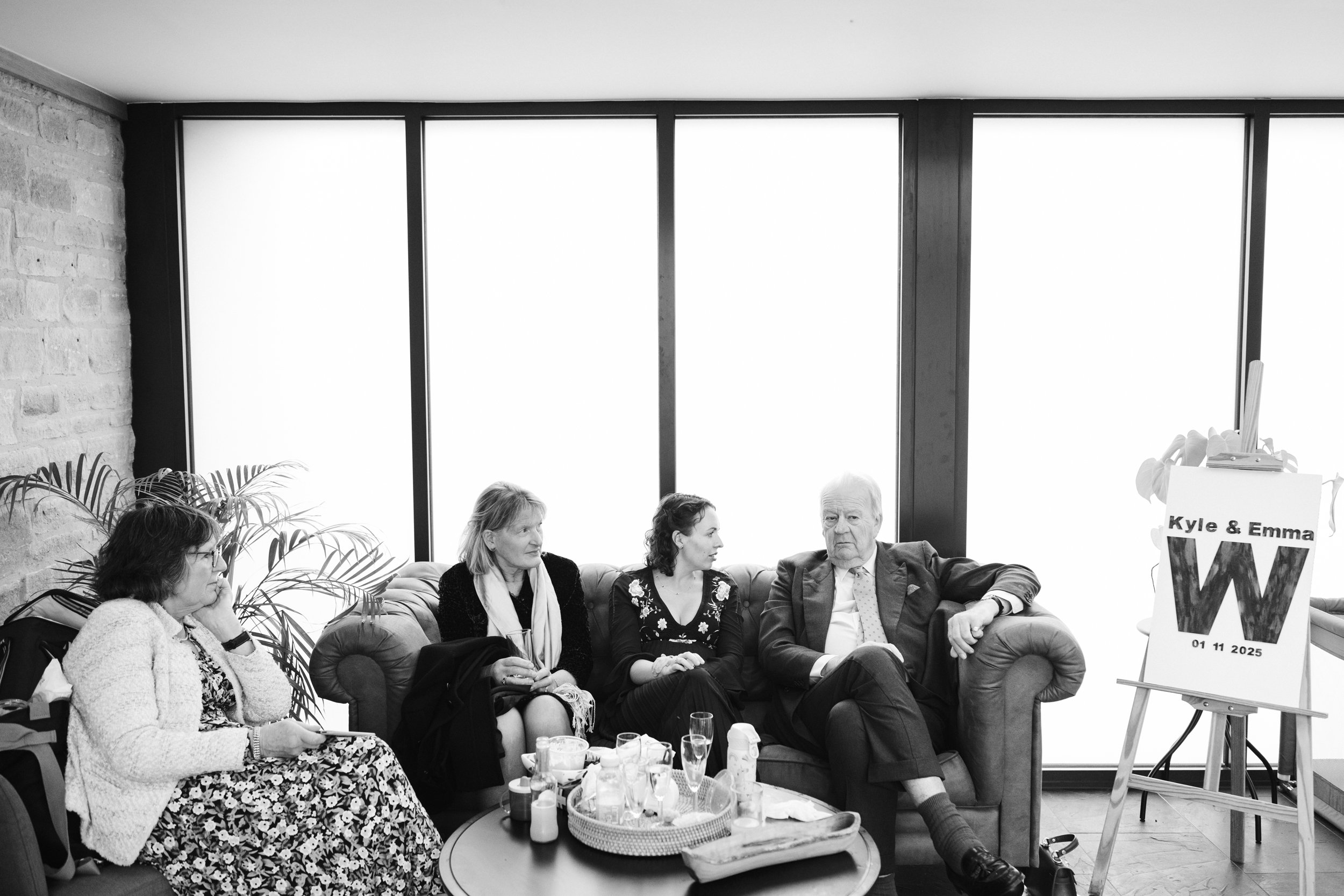 Four people sitting on a couch in a room with large windows, beside a table with drinks and snacks, and a sign on an easel reading 'Kyle & Emma W, 01 11 2025'.