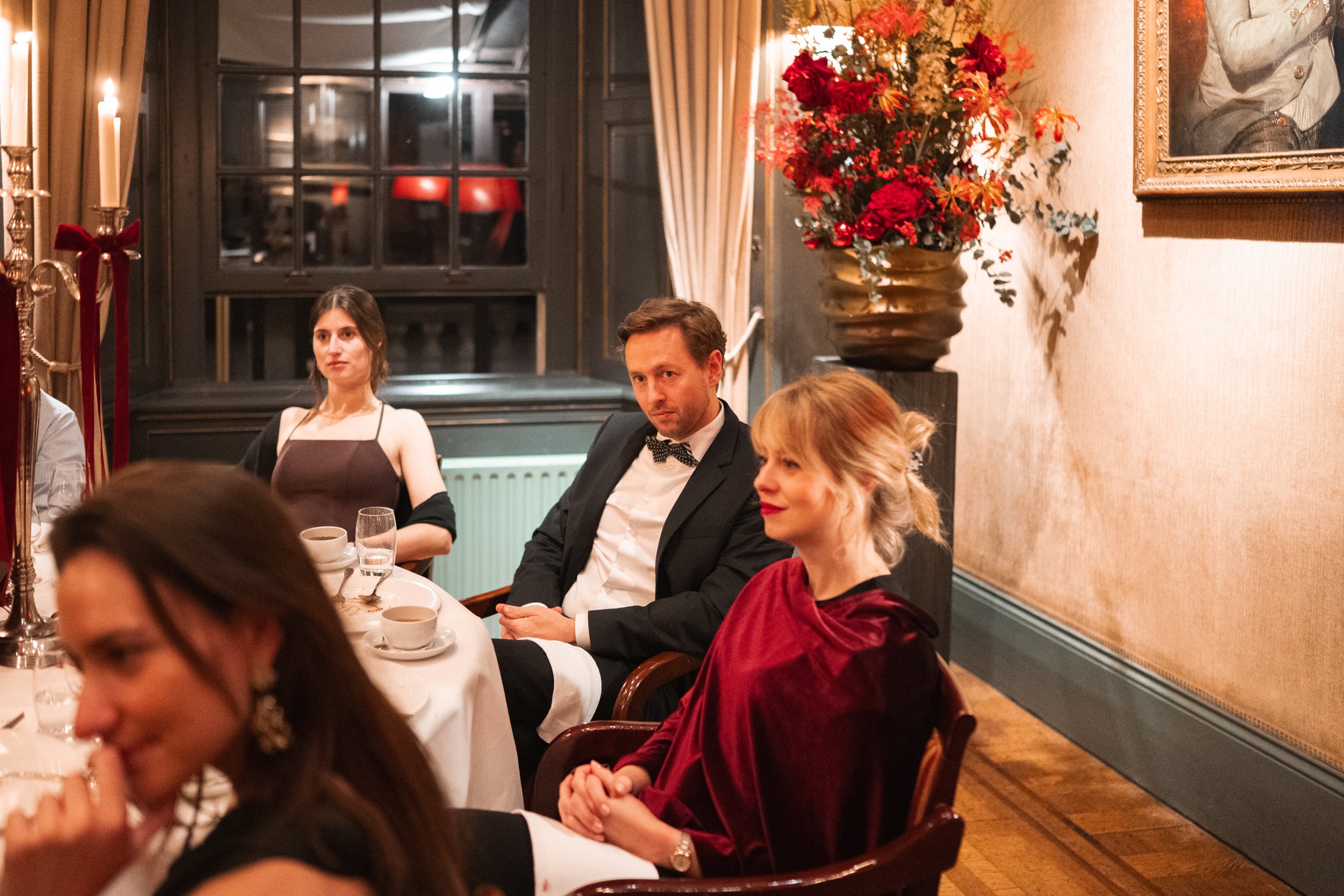 People sitting at a formal dinner table in a decorated, elegant room with flowers, curtains, and framed art on the wall.