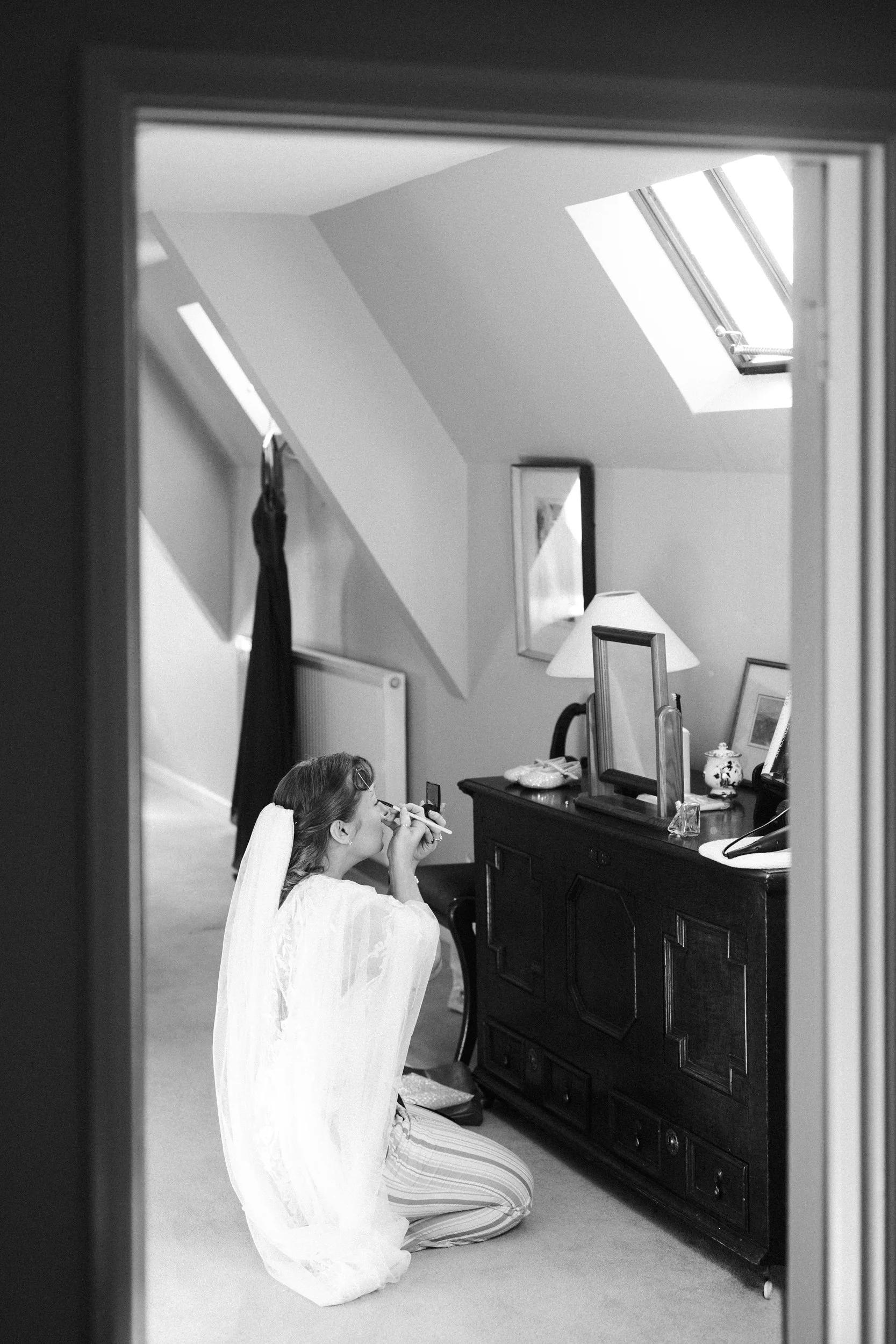 A woman sitting on the floor applying makeup while looking into a small mirror, in a room with sloped ceilings and a skylight.
