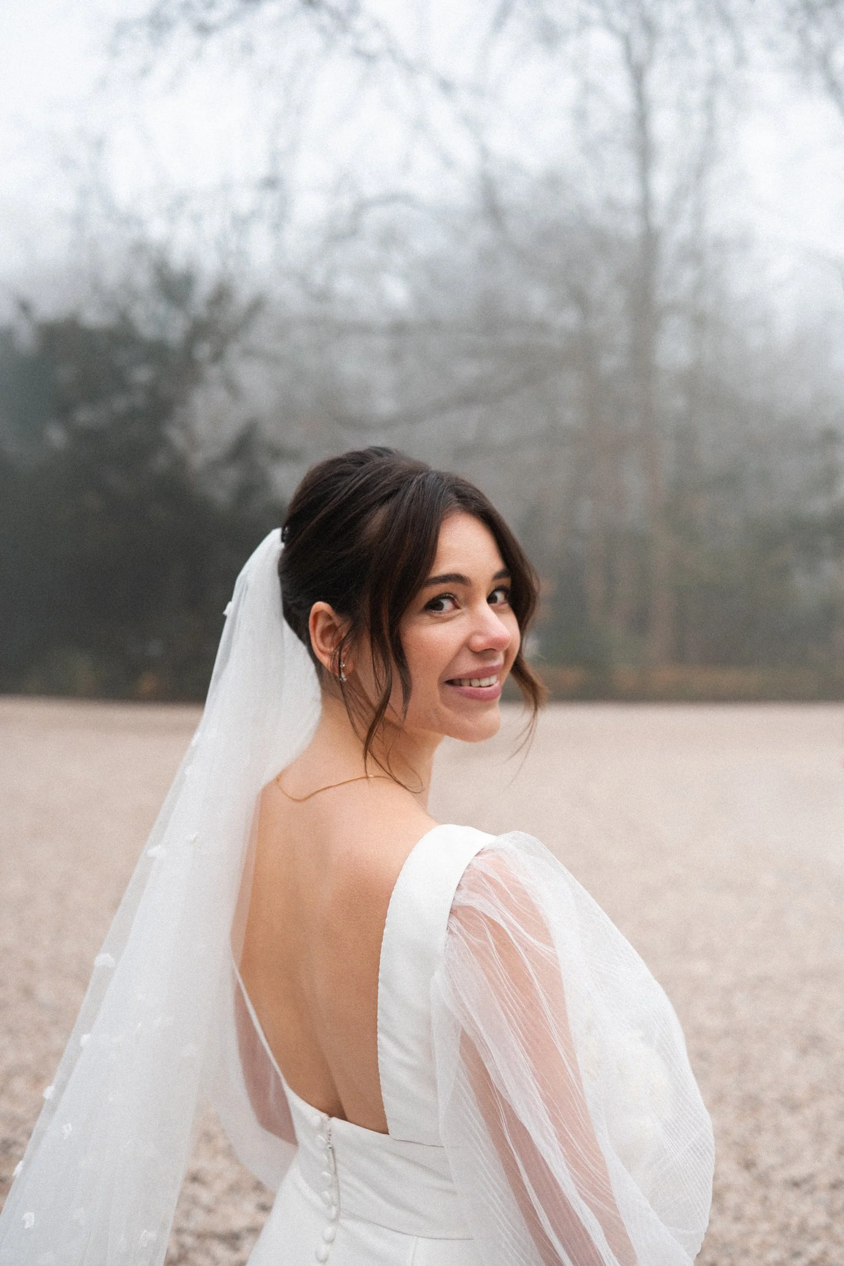 A smiling bride in a white wedding dress with sheer puffy sleeves and a veil, standing outdoors with a blurred background of leafless trees and a misty sky.