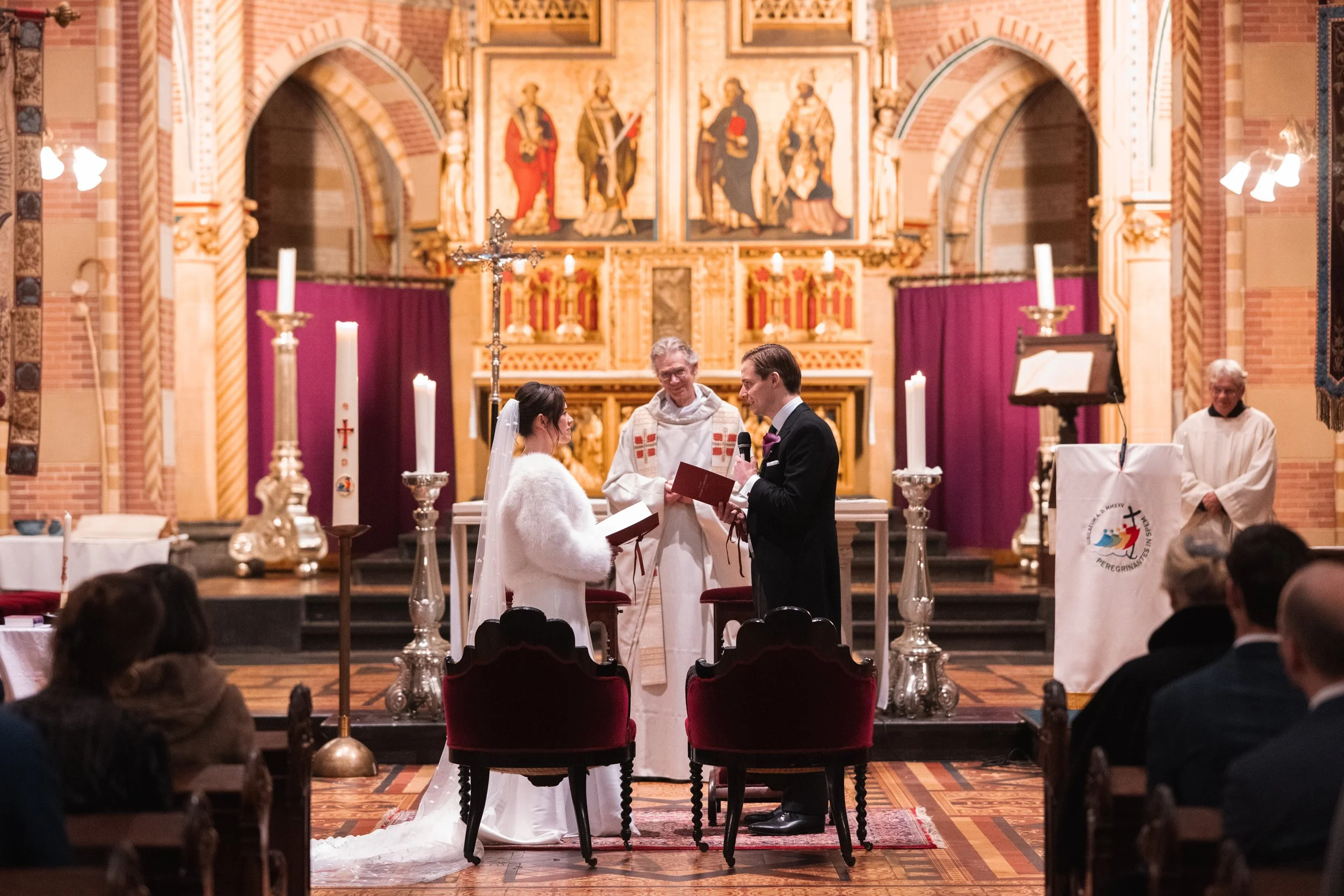 A wedding ceremony taking place inside a church with a priest, bride, and groom standing at the altar. The bride is wearing a white dress and veil, while the groom is dressed in a black tuxedo. There are candles and religious artwork in the backgroun