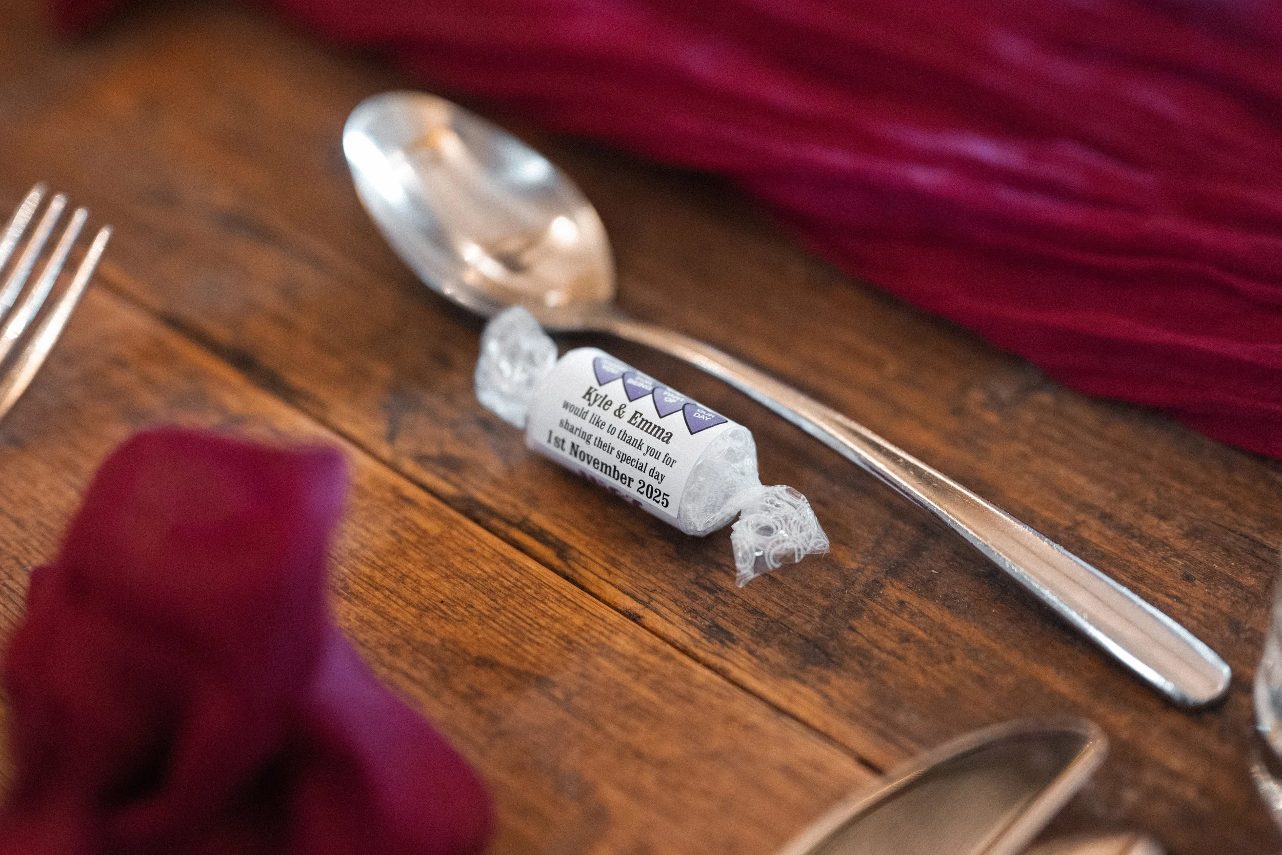 A personalized wrapped candy on a wooden table with a silver spoon and a fork nearby, pink cloth in the background, and a person's hand in a red glove is partially visible in the lower left corner.