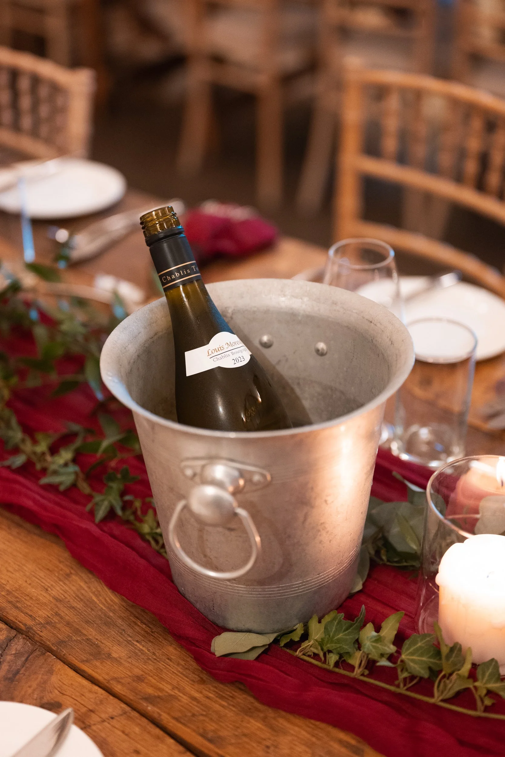 A bottle of red wine in an ice bucket on a dining table, with table settings and candles in the background.