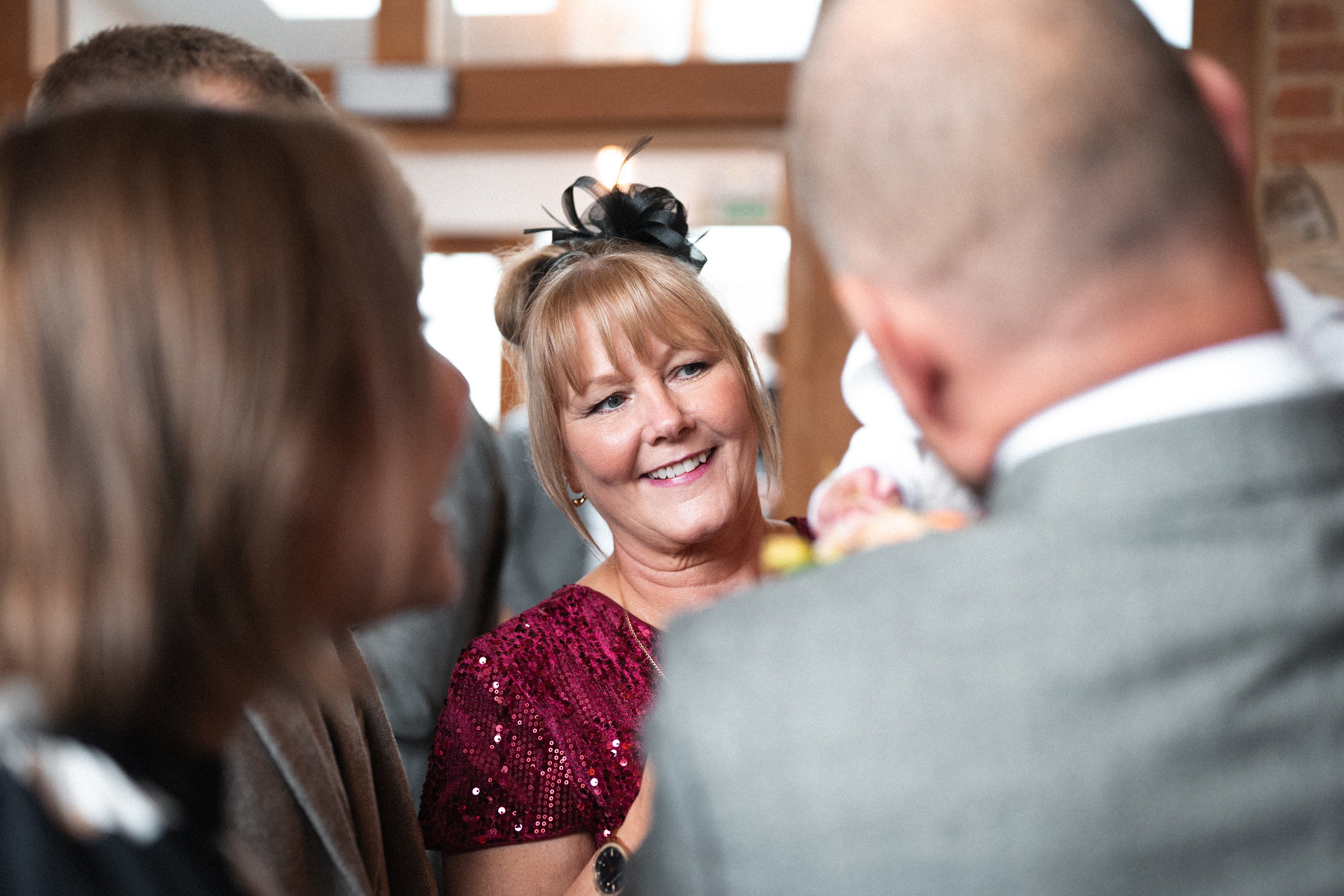 A woman with red hair wearing a red sequined dress smiling and talking to a group of people at what appears to be a social gathering or celebration.