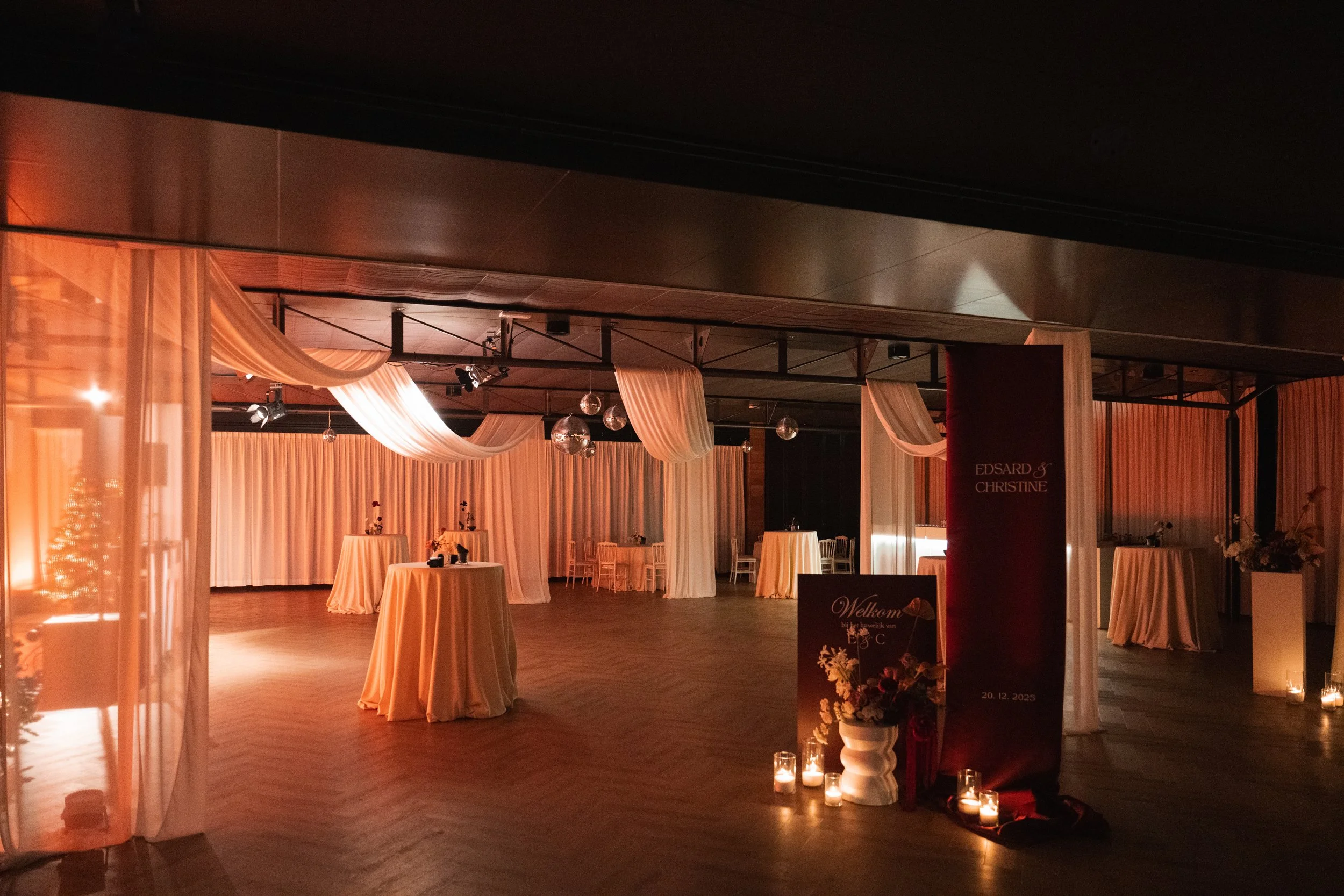 An elegantly decorated event space with tall tables draped in peach-colored tablecloths, sheer curtains hanging from the ceiling, soft warm lighting, and floral arrangements. Candles are placed on the floor, enhancing the cozy atmosphere. A sign disp