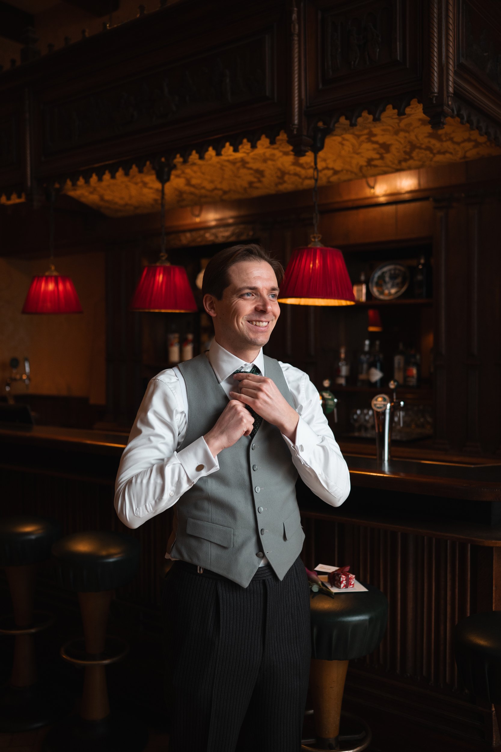 A smiling man in a white shirt, gray vest, and dark trousers adjusting his tie inside a dimly lit bar with red lamps and wooden decor.