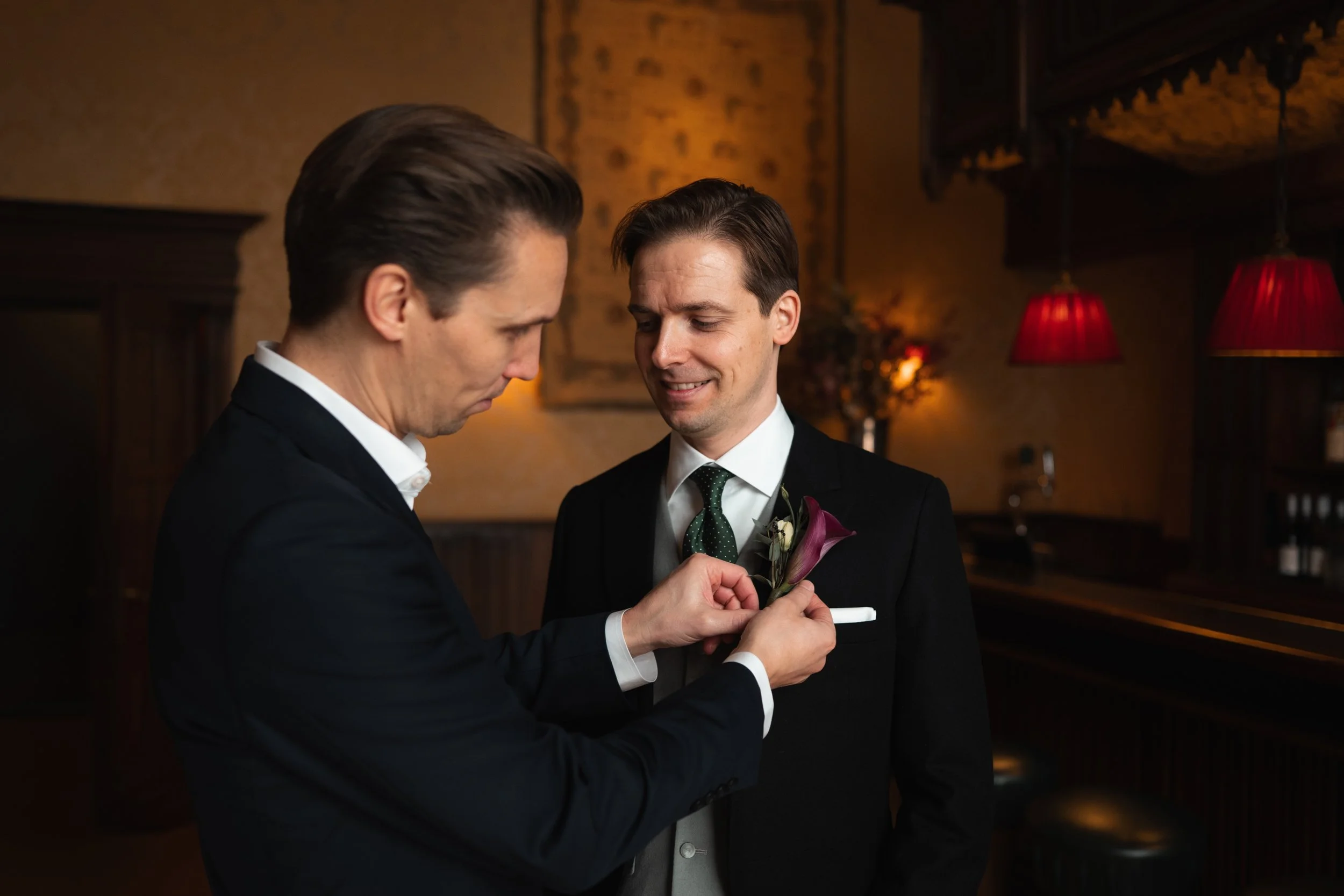 Two men in suits, one placing a boutonniere on the other's jacket in a warmly lit restaurant interior.