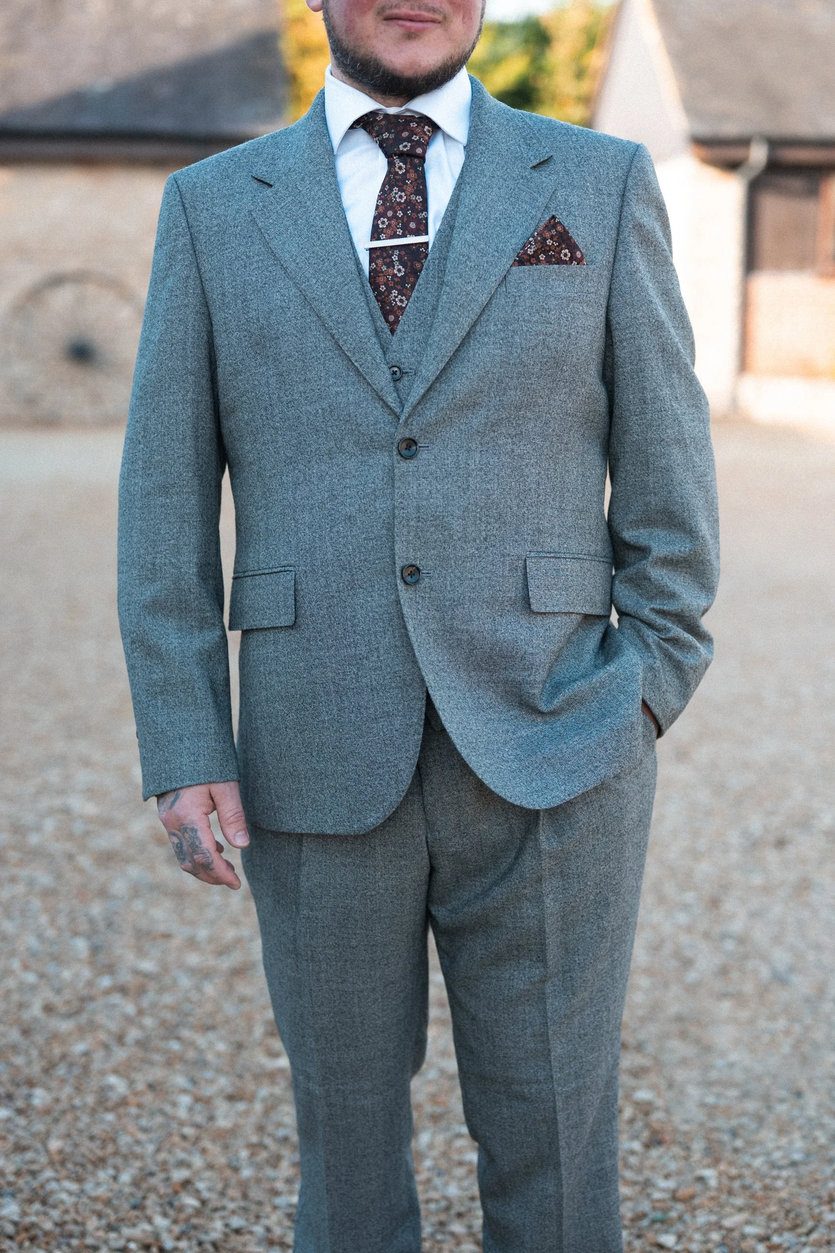 Close-up of a man wearing a gray suit, white shirt, and the top of a patterned tie, standing outdoors in front of a rustic house with brick and stone walls.