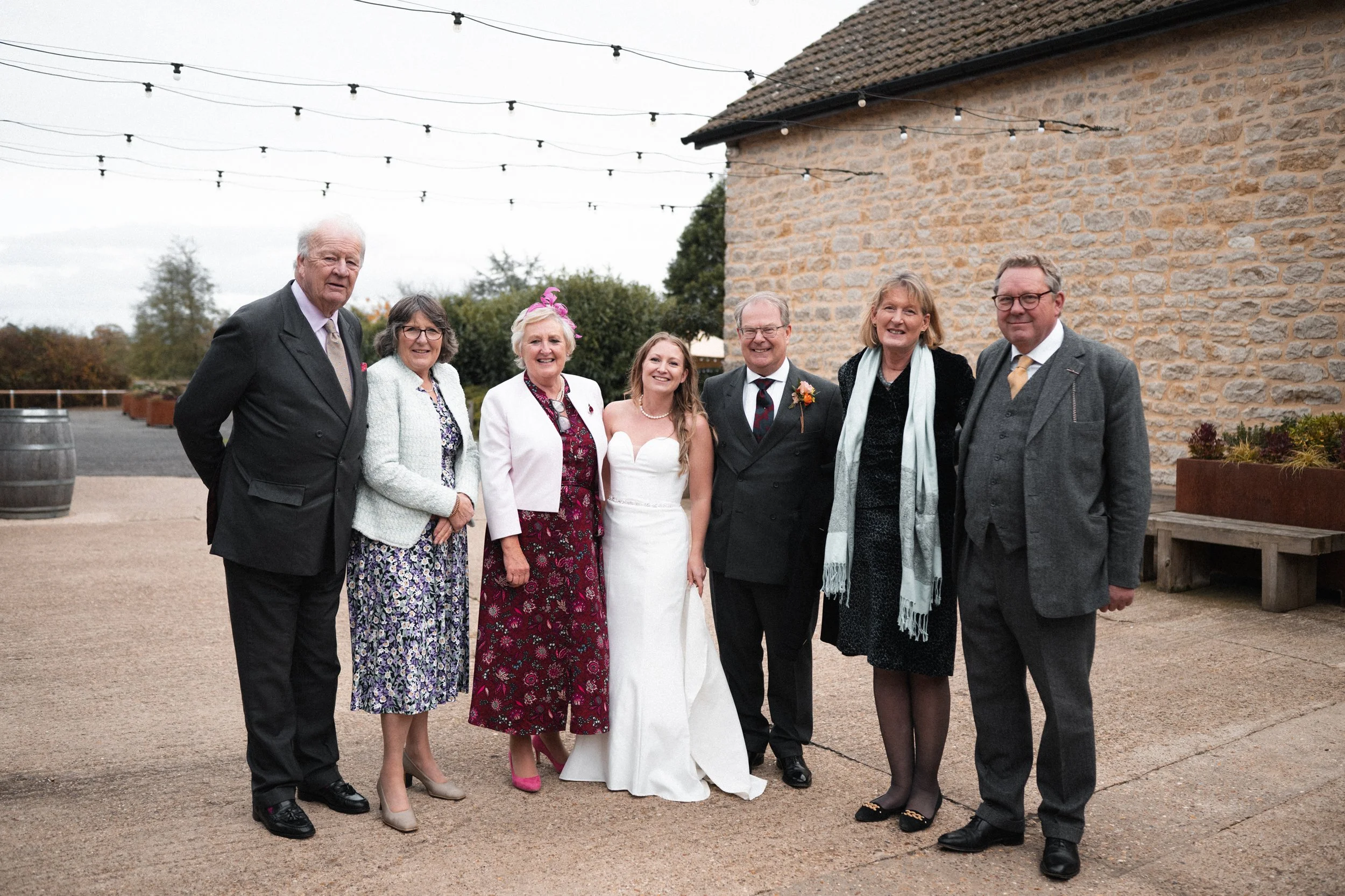 Group of eight people, including bride in white wedding dress and groom in black suit, standing outdoors near a stone building with string lights overhead, celebrating a wedding.