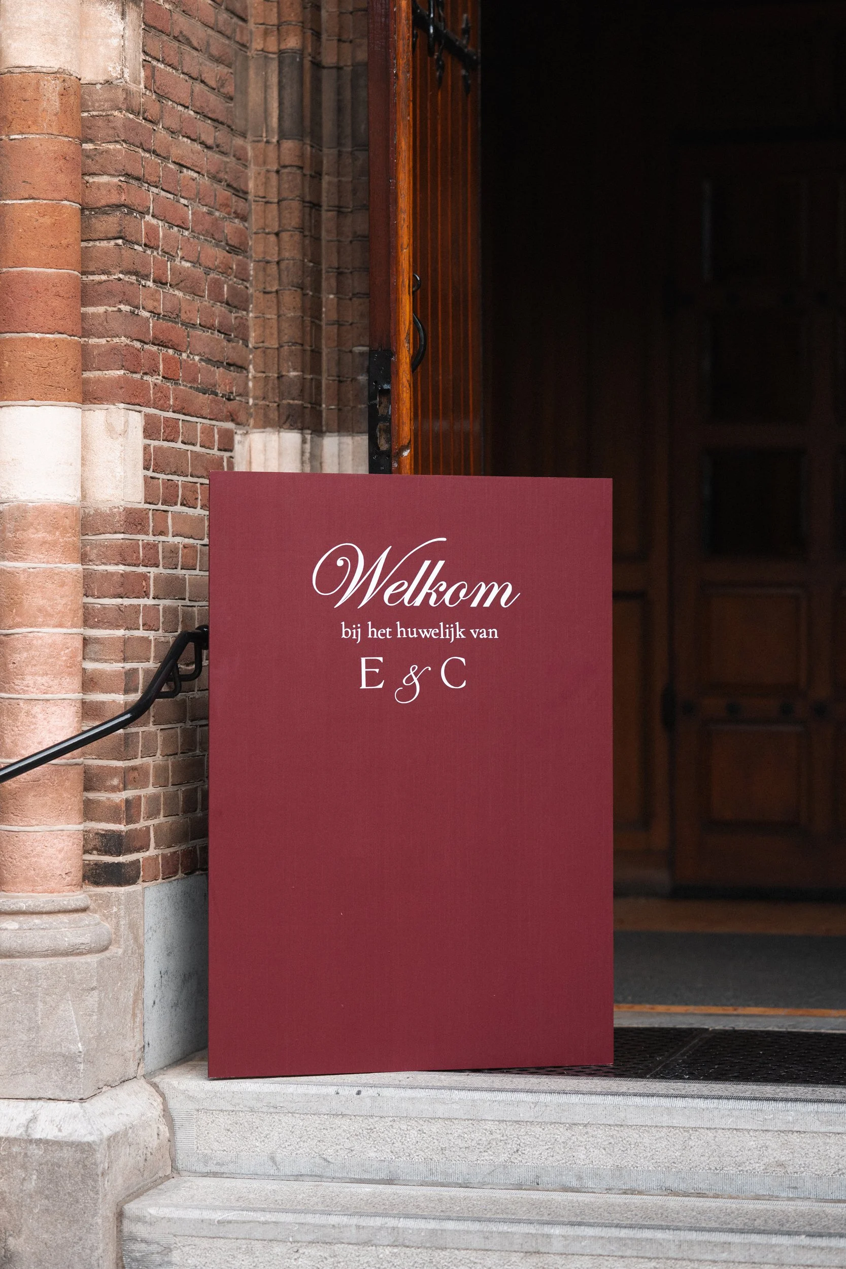 A maroon wedding sign with white cursive and serif text reading 'Welkom bij het huwelijk van E & C,' placed outside a brick church entrance.