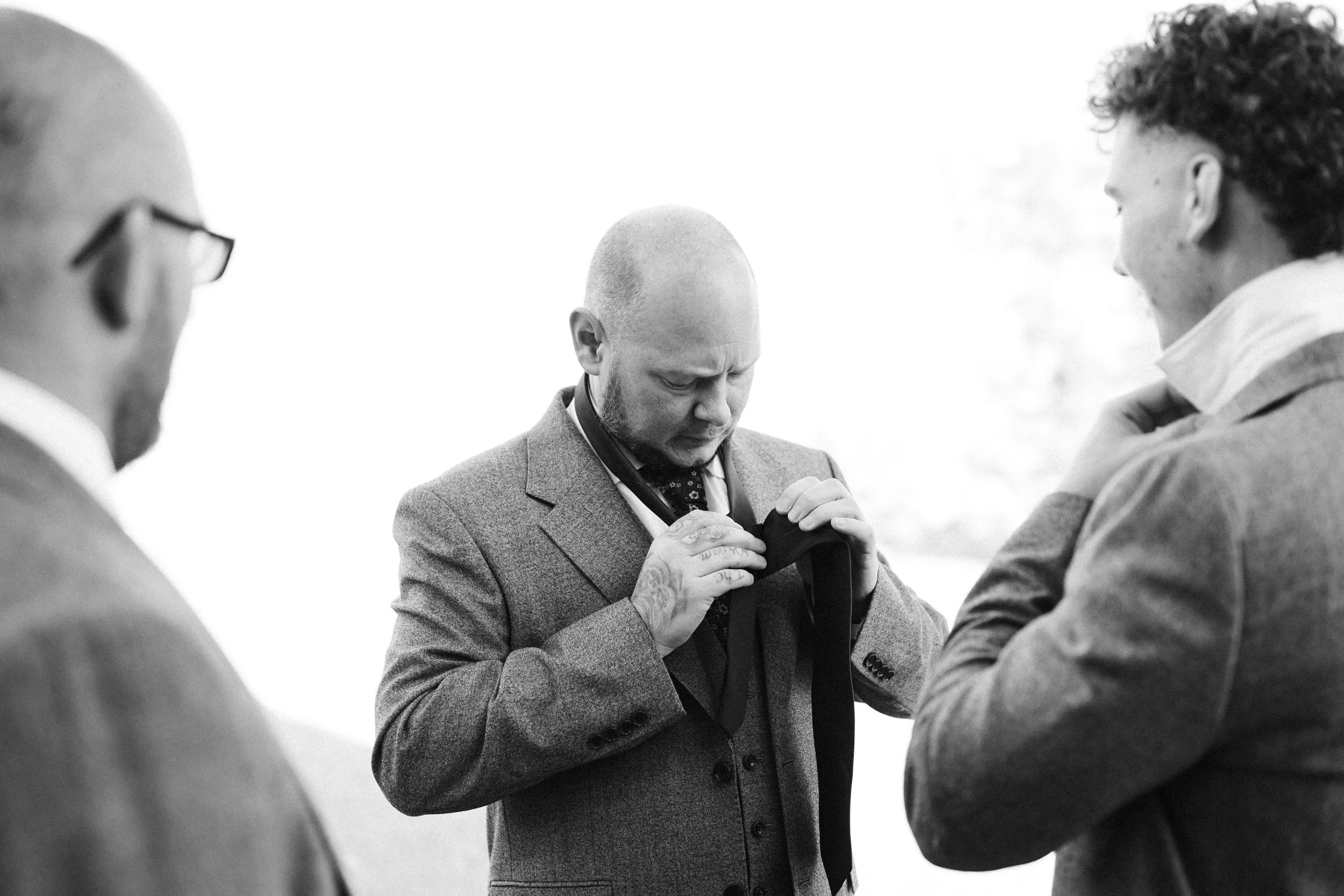 Three men in suits preparing for a formal event, possibly a wedding, with one adjusting a tie.